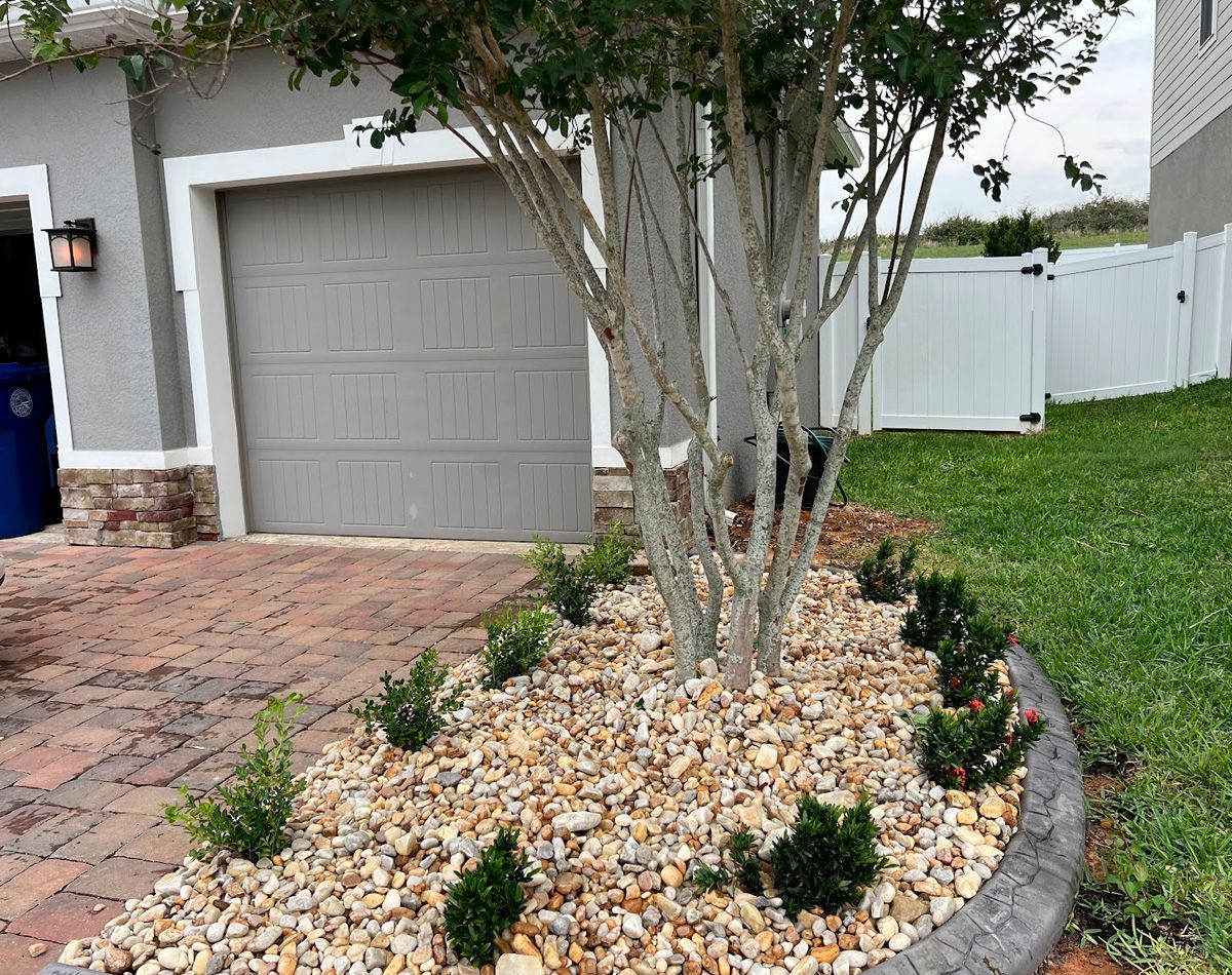 Landscaped yard with a tree and shrubs, gravel bed, and brick driveway next to a garage.