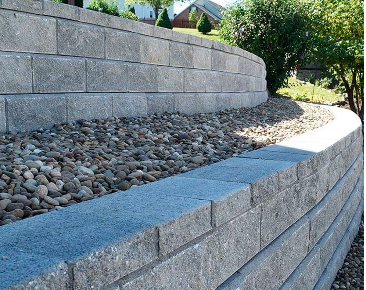 Gray retaining wall made of rectangular blocks, holding back gravel and a hillside.