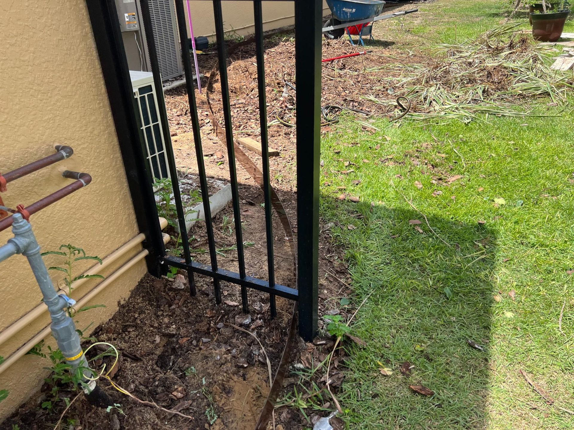 Black metal gate next to beige building, gas pipes, and green grass. Dirt and debris are present.