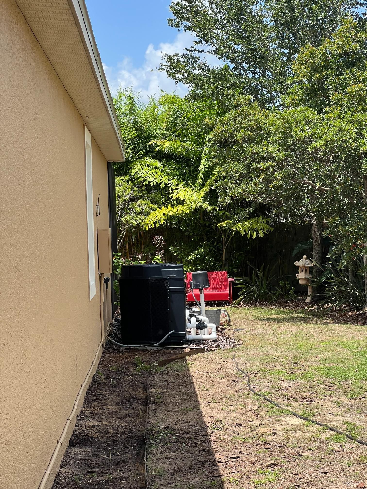 Side of a beige house with a black trash can and an air conditioning unit; a backyard with trees.