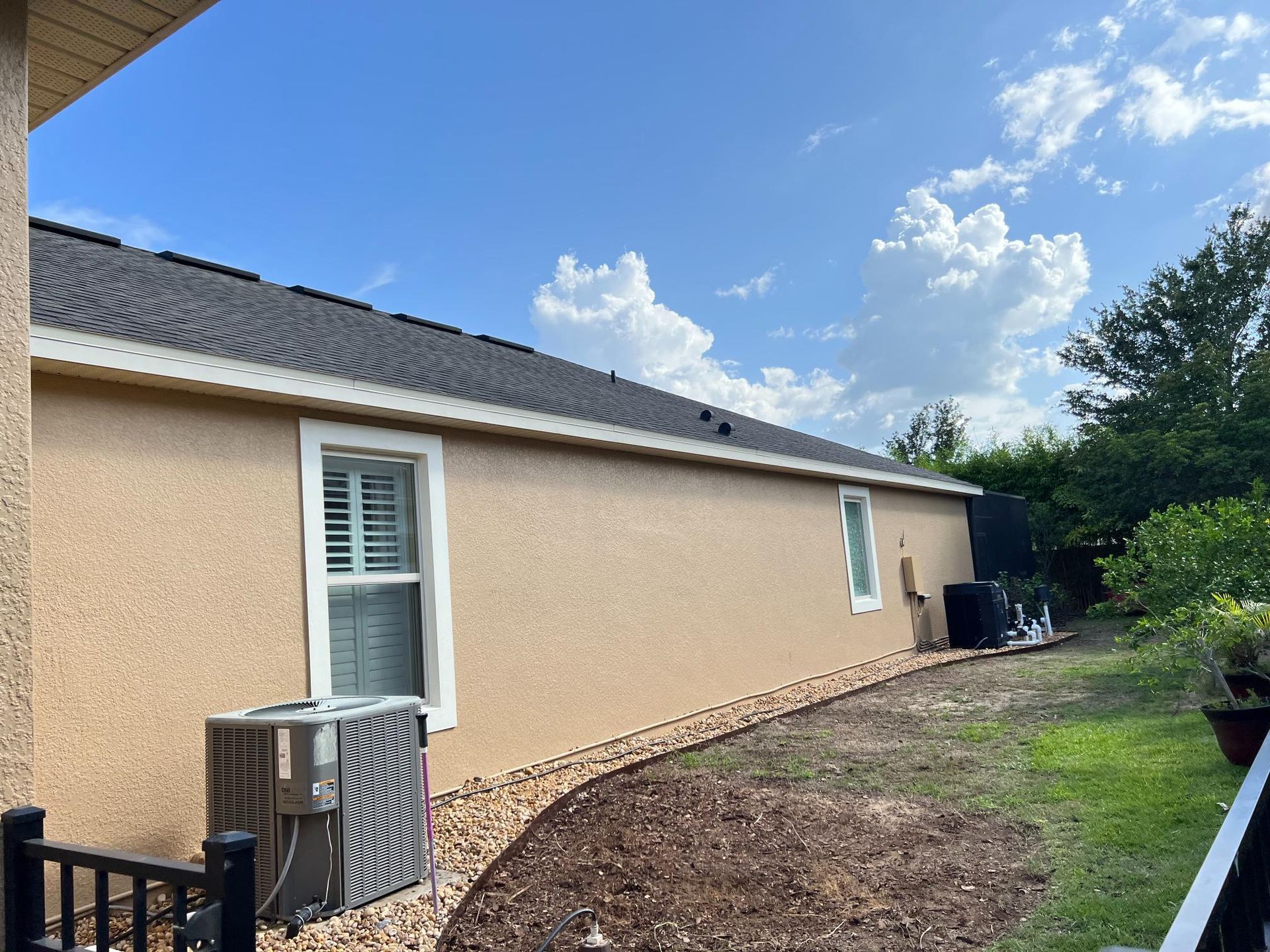 Tan stucco house exterior with dark roof, windows, and air conditioning units.