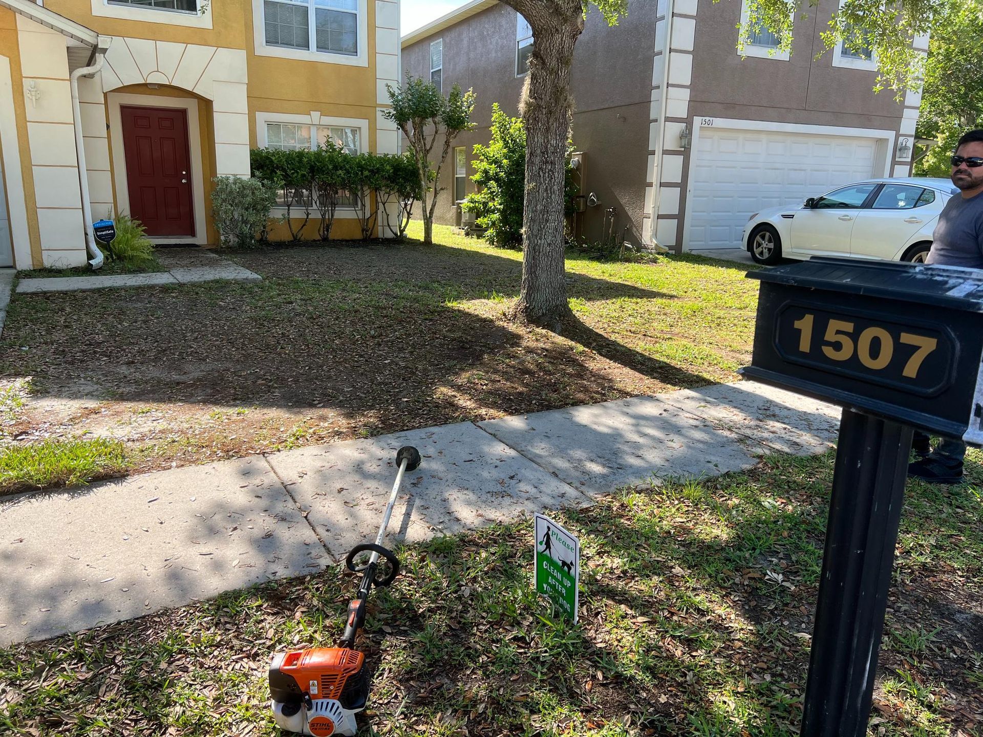 A lawn being trimmed in front of a house. A string trimmer rests on the ground next to a mailbox.