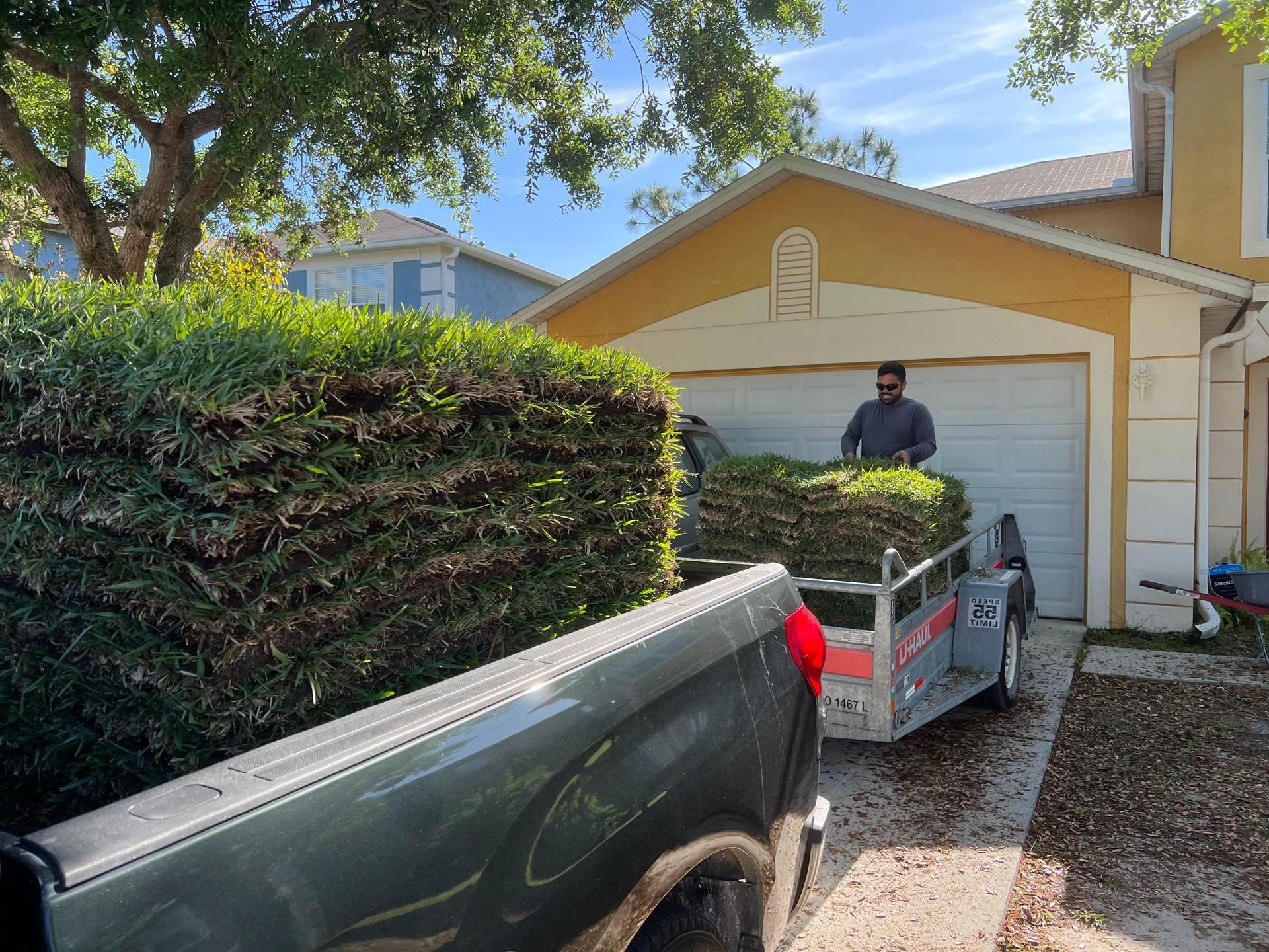 Truck with trailer loaded with cut bushes; person loading more in driveway of yellow house.