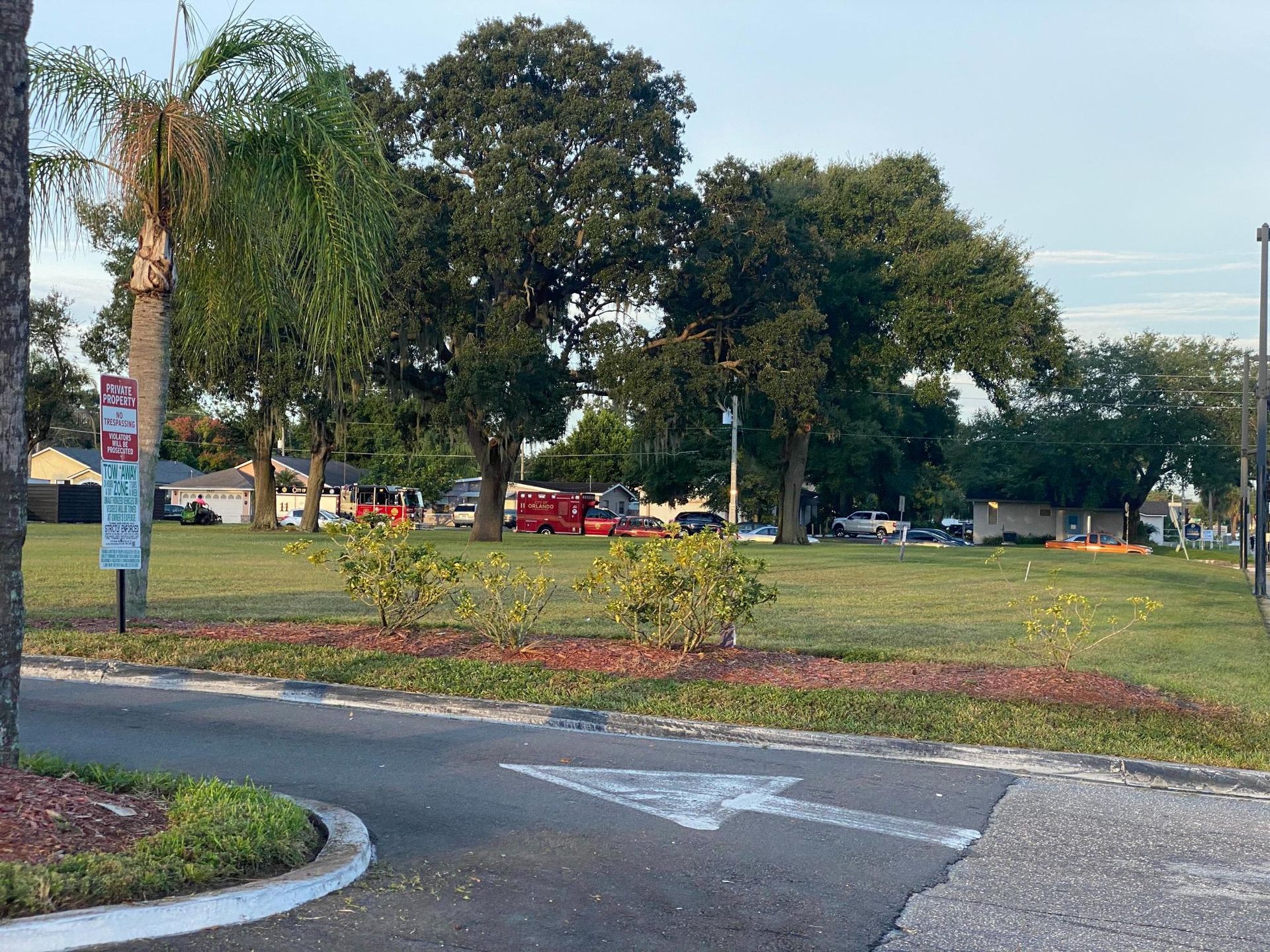 Grassy park area with trees and shrubs. Paved path with an arrow. Parked vehicles visible in the distance.
