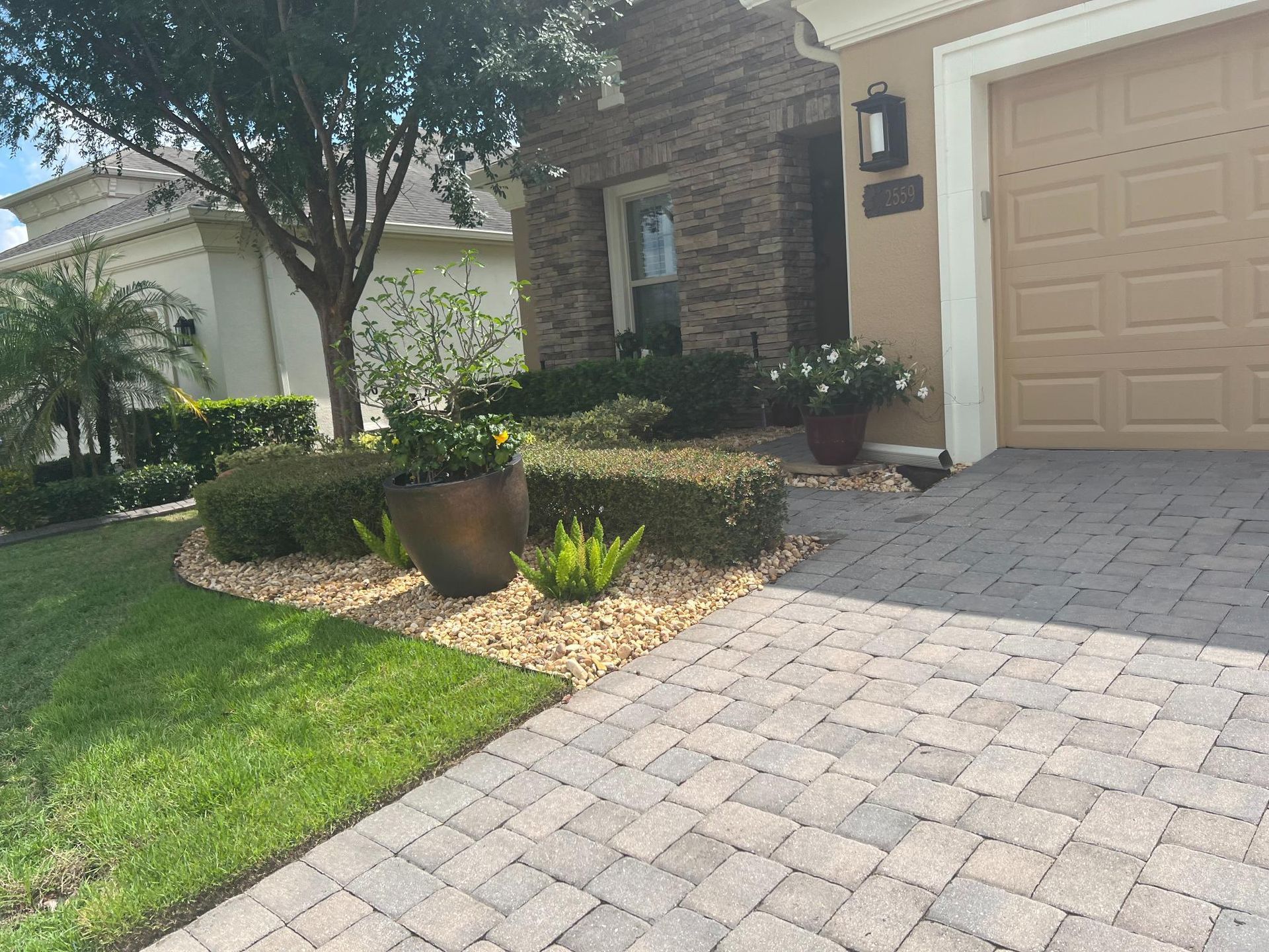Brick paved driveway leads to a home with well-manicured landscaping, including shrubs and flower pots.