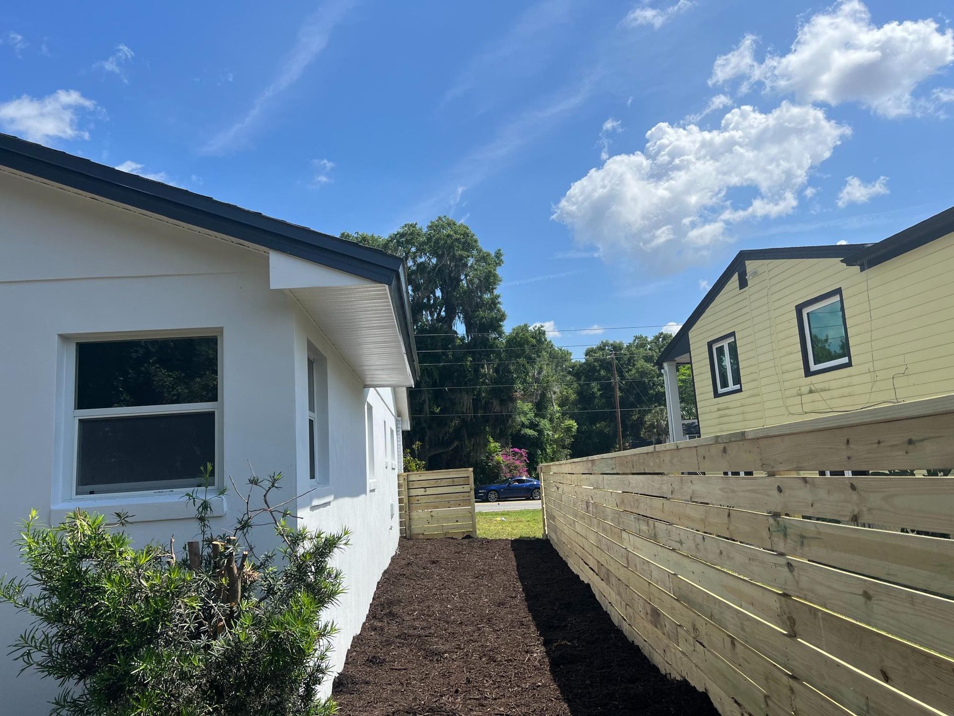A narrow walkway between two houses. One is white, the other yellow, separated by a wooden fence, under a partly cloudy sky.