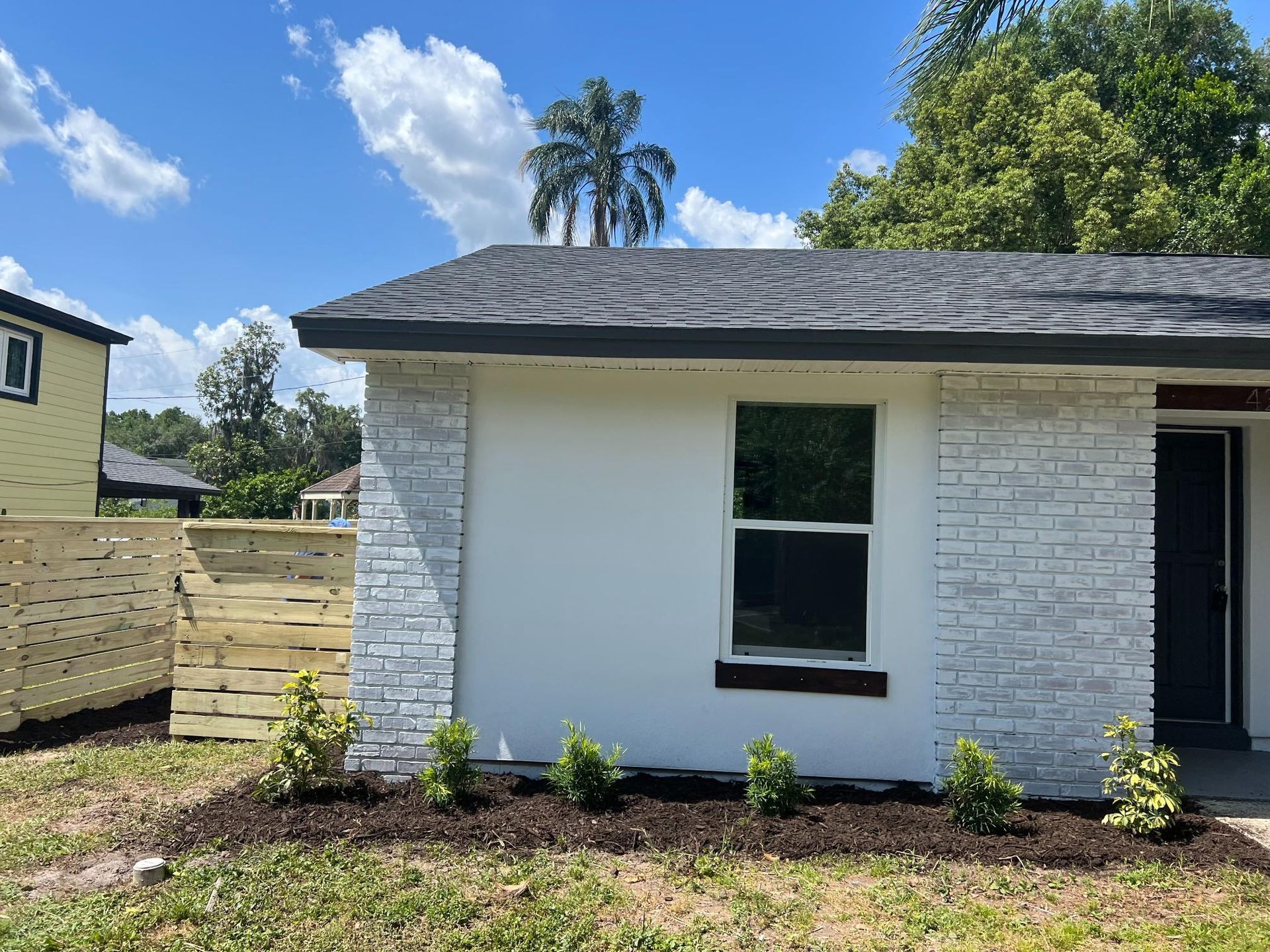 White house with dark roof and brick accents, window, plants, and wooden fence.
