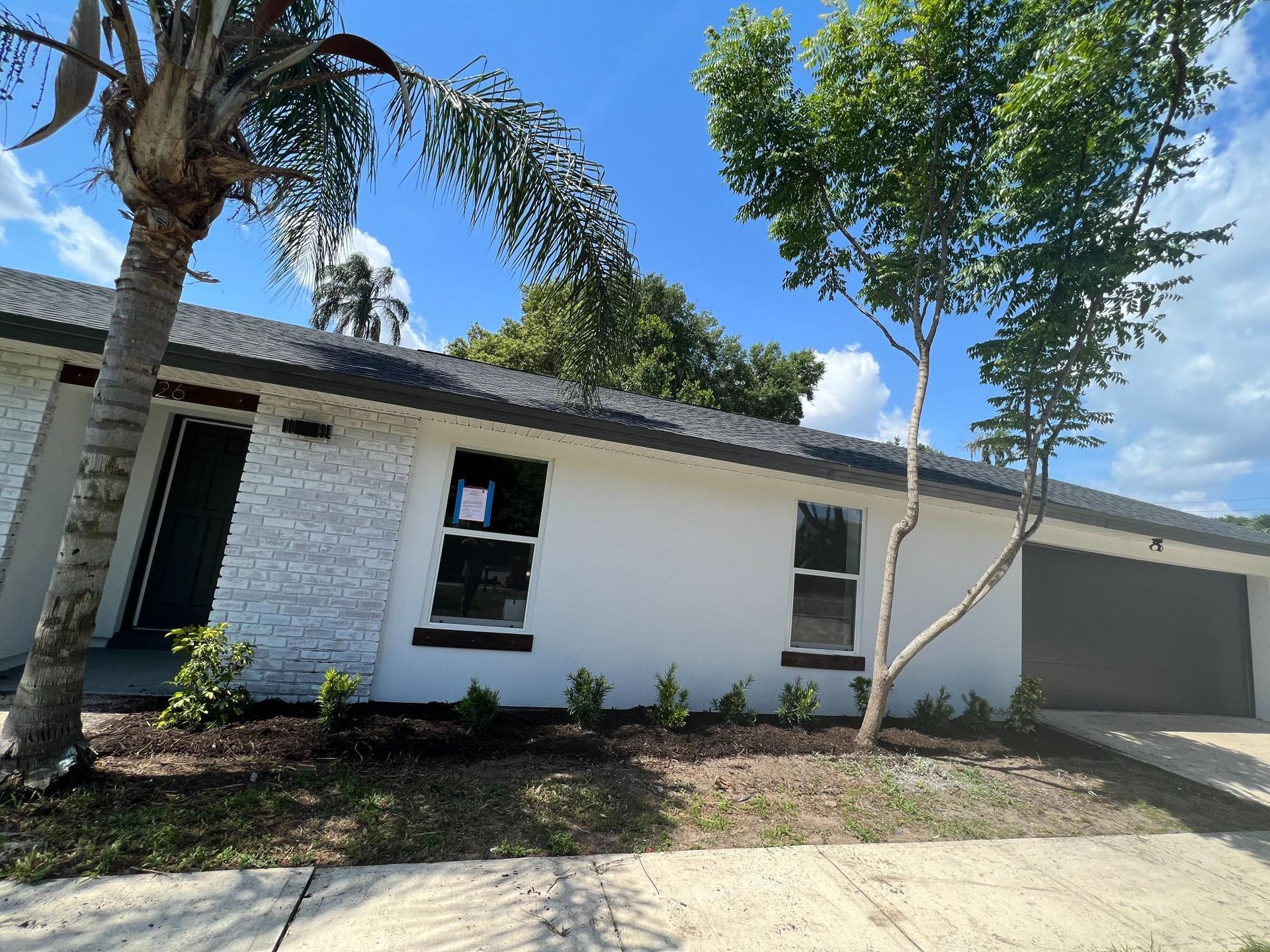 White house with black trim, windows, and garage door. Palm and tree in front, blue sky.
