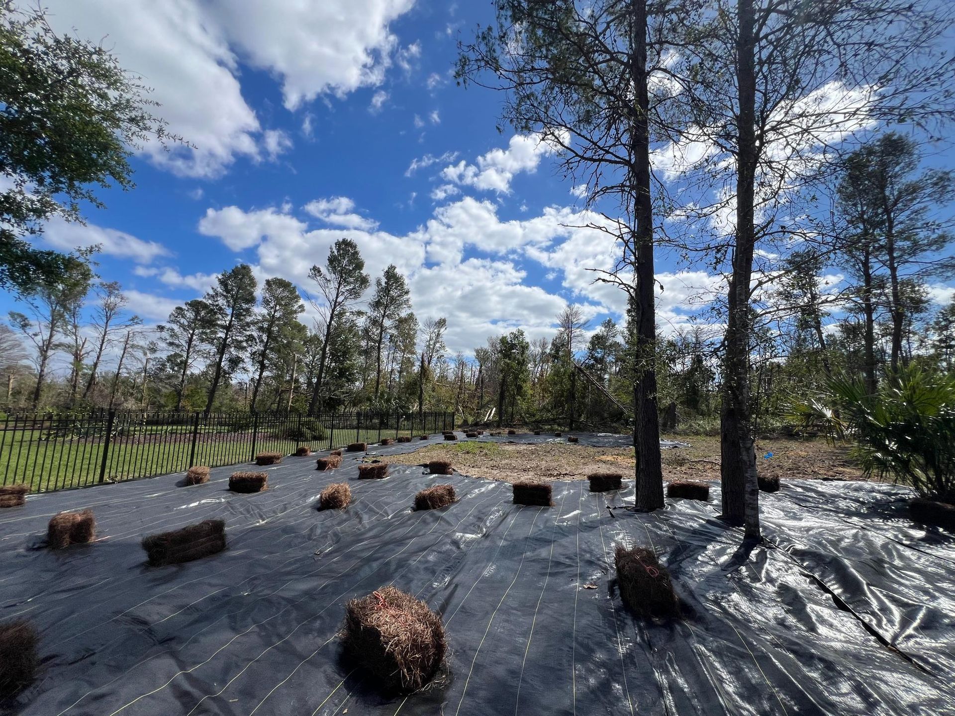 Field with dark tarp, straw bales, and trees under a blue sky with clouds.