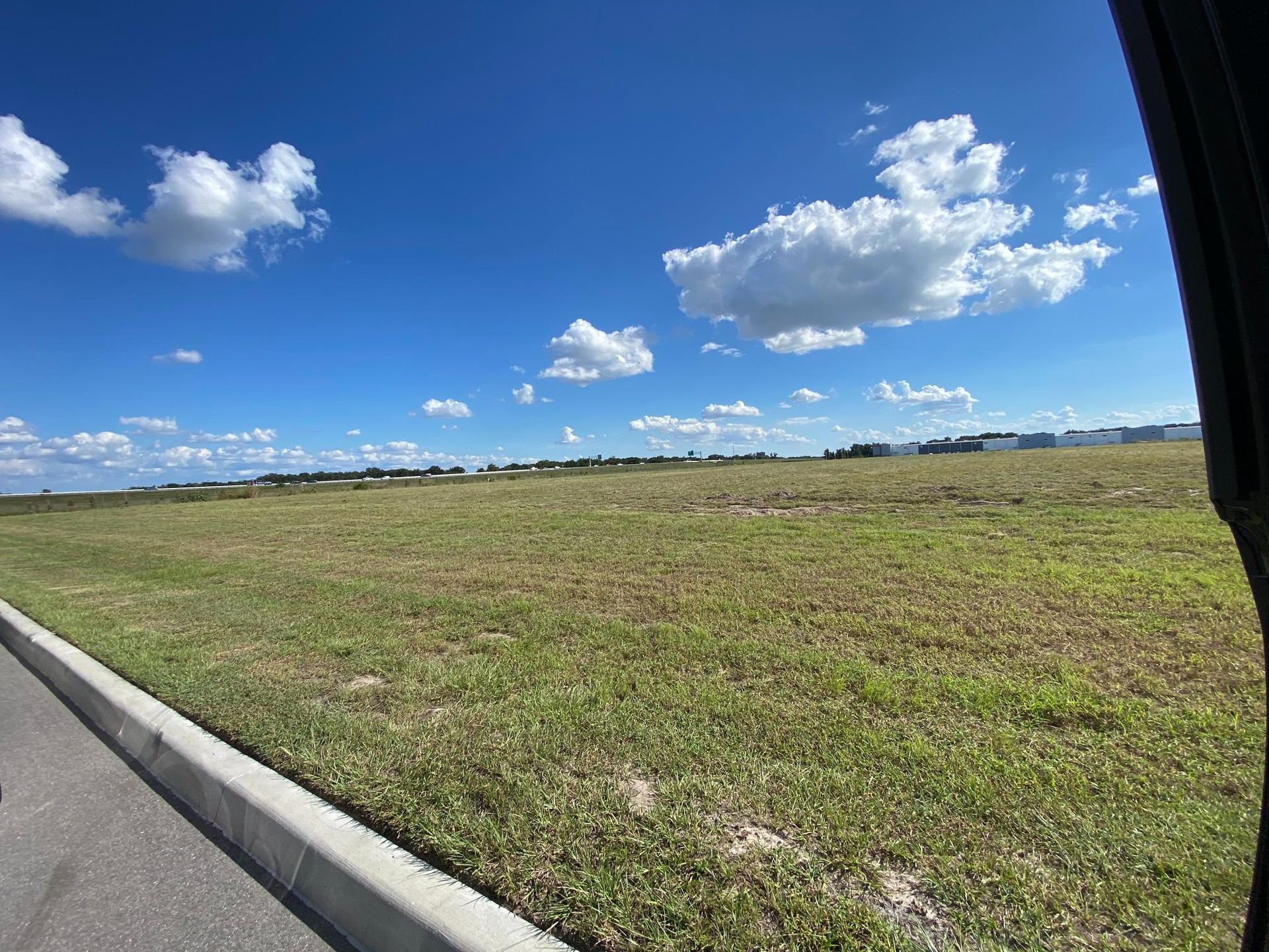 Grassy field under a blue sky with scattered clouds, next to a paved road.
