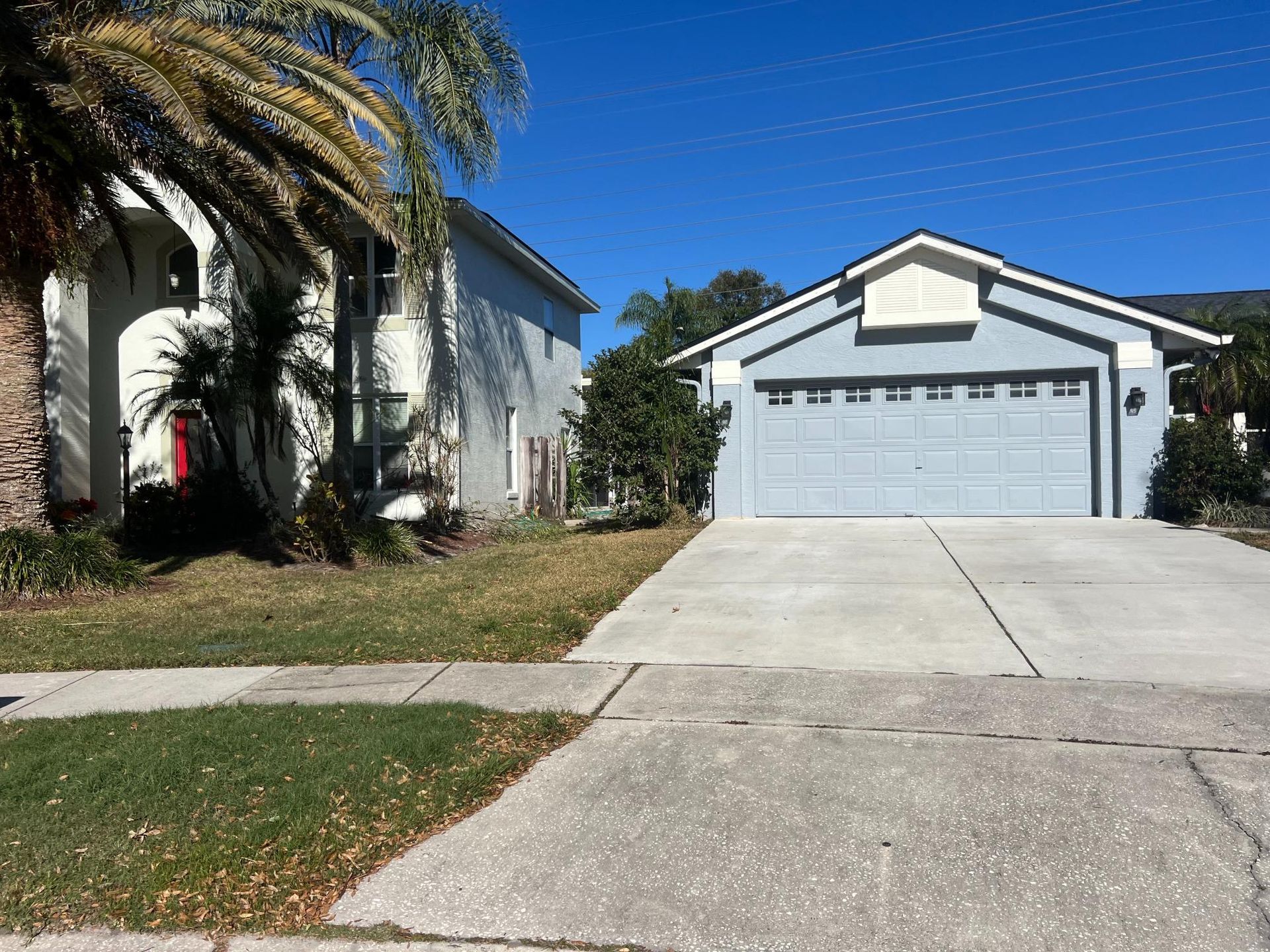 Two-story light blue house with a two-car garage. A driveway leads to the street with a grassy yard.