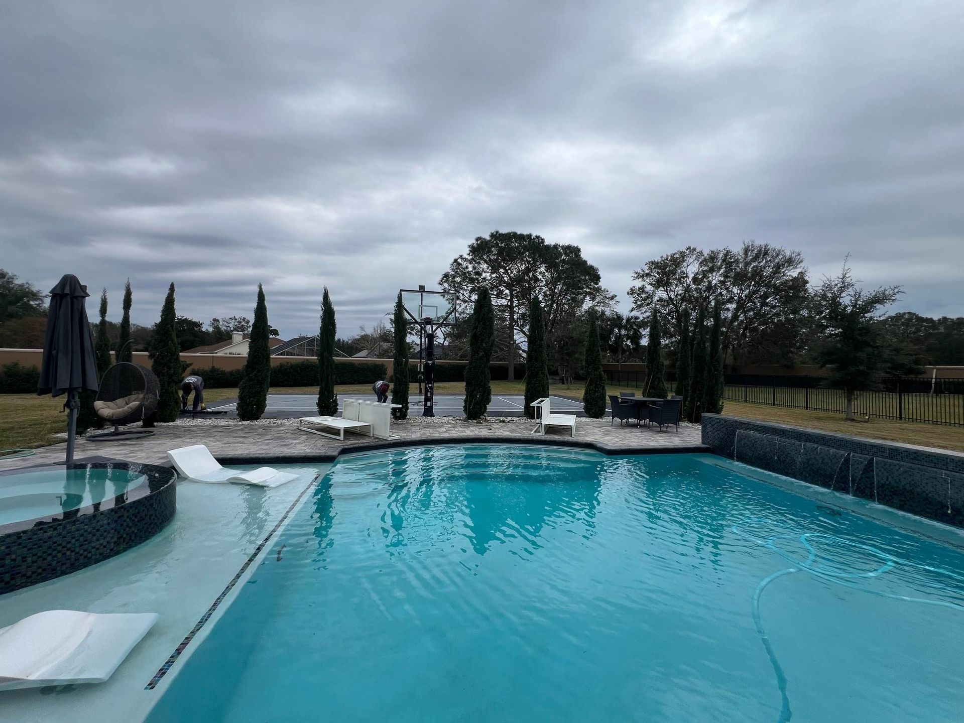 Pool with blue water and tiled edges, surrounded by lounge chairs, trees, and cloudy sky.