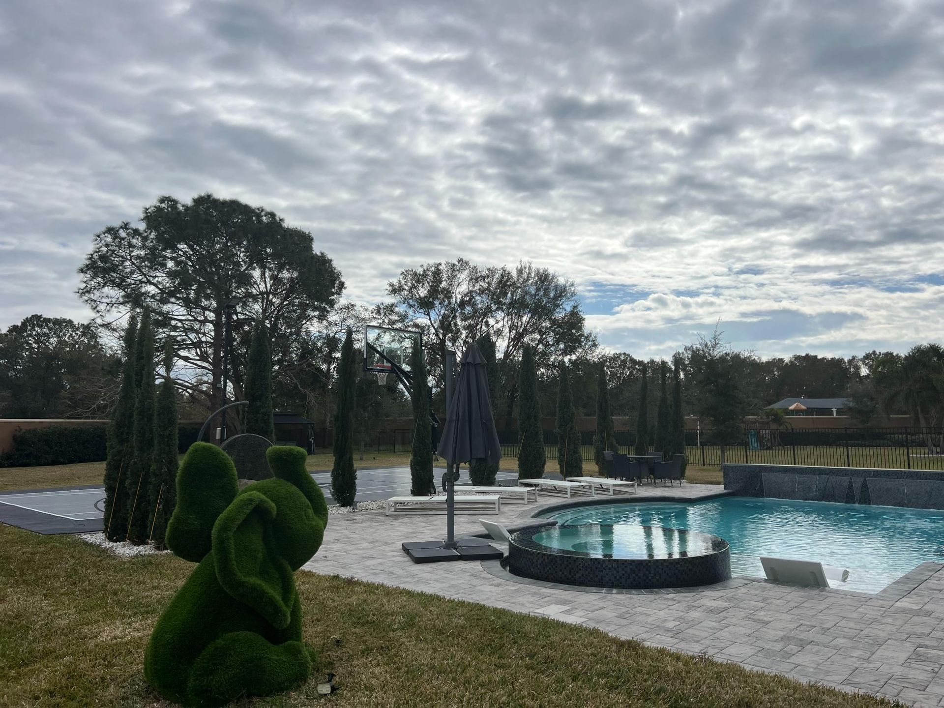 Backyard pool with white stone patio, elephant topiary, and cloudy sky.