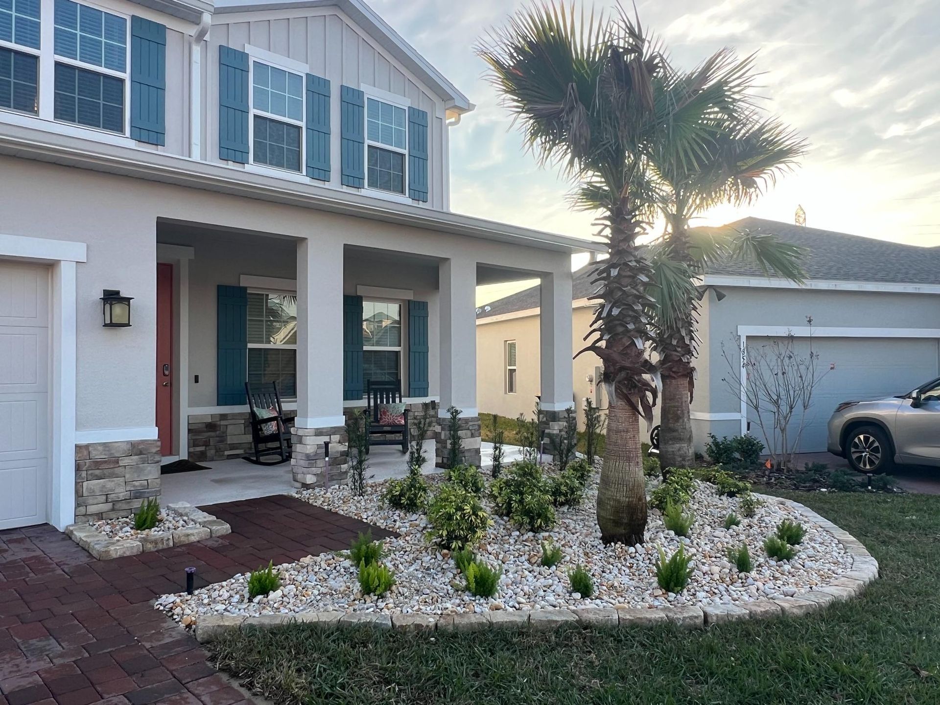 Two-story house with a porch and landscaped yard. Light gray paint, blue shutters, and palm trees.