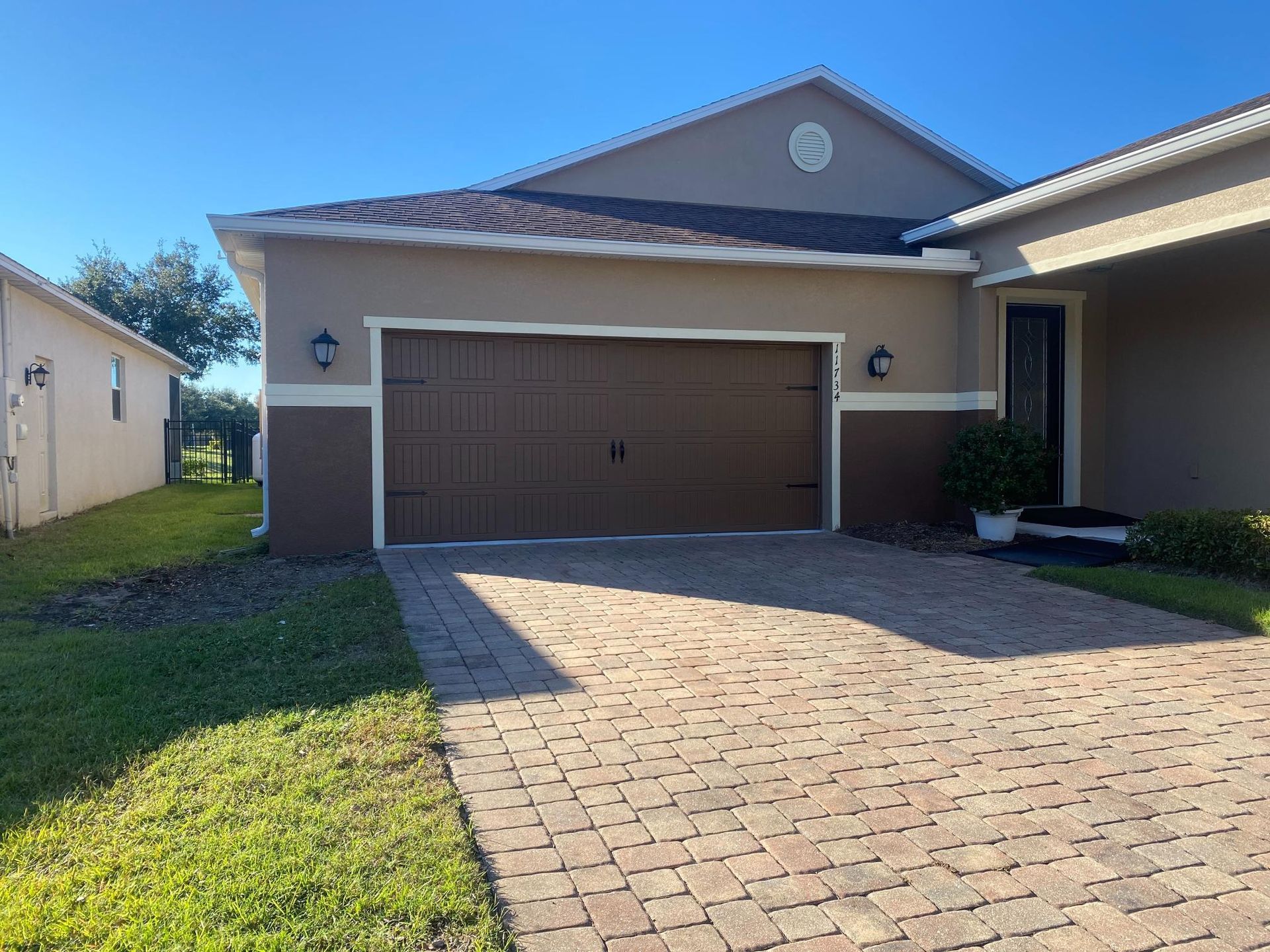 Tan-colored house with brown garage door, paved driveway, and green lawn under a blue sky.