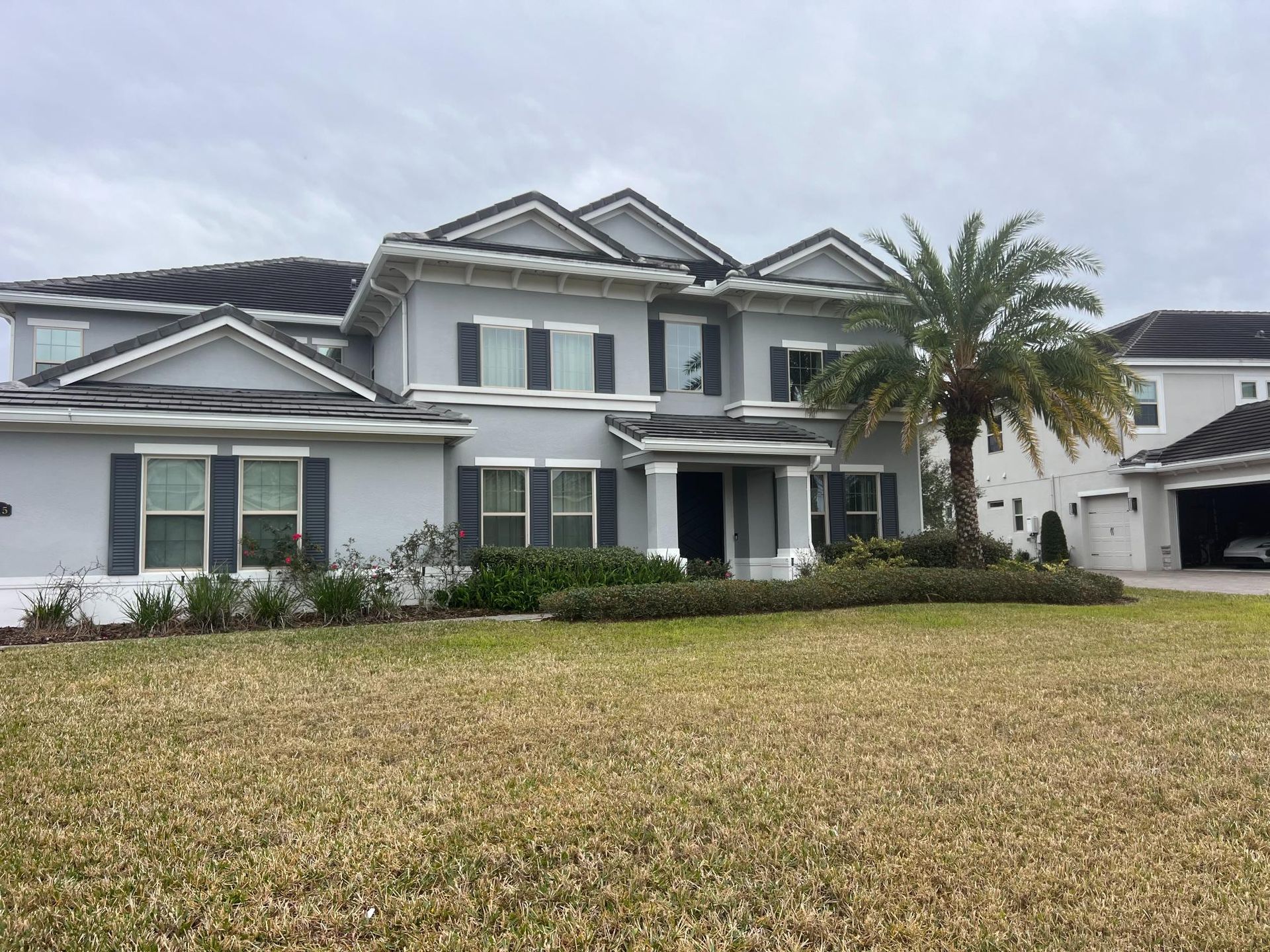 Two-story gray house with black shutters, palm tree, and brown grass lawn under cloudy sky.