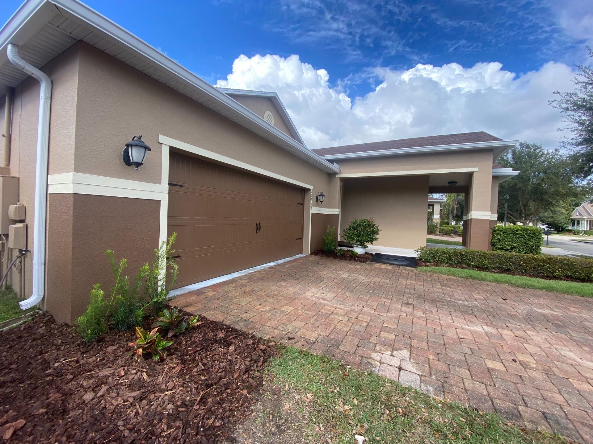 Tan and brown house with a brick driveway and a brown garage door. Blue sky with white clouds.