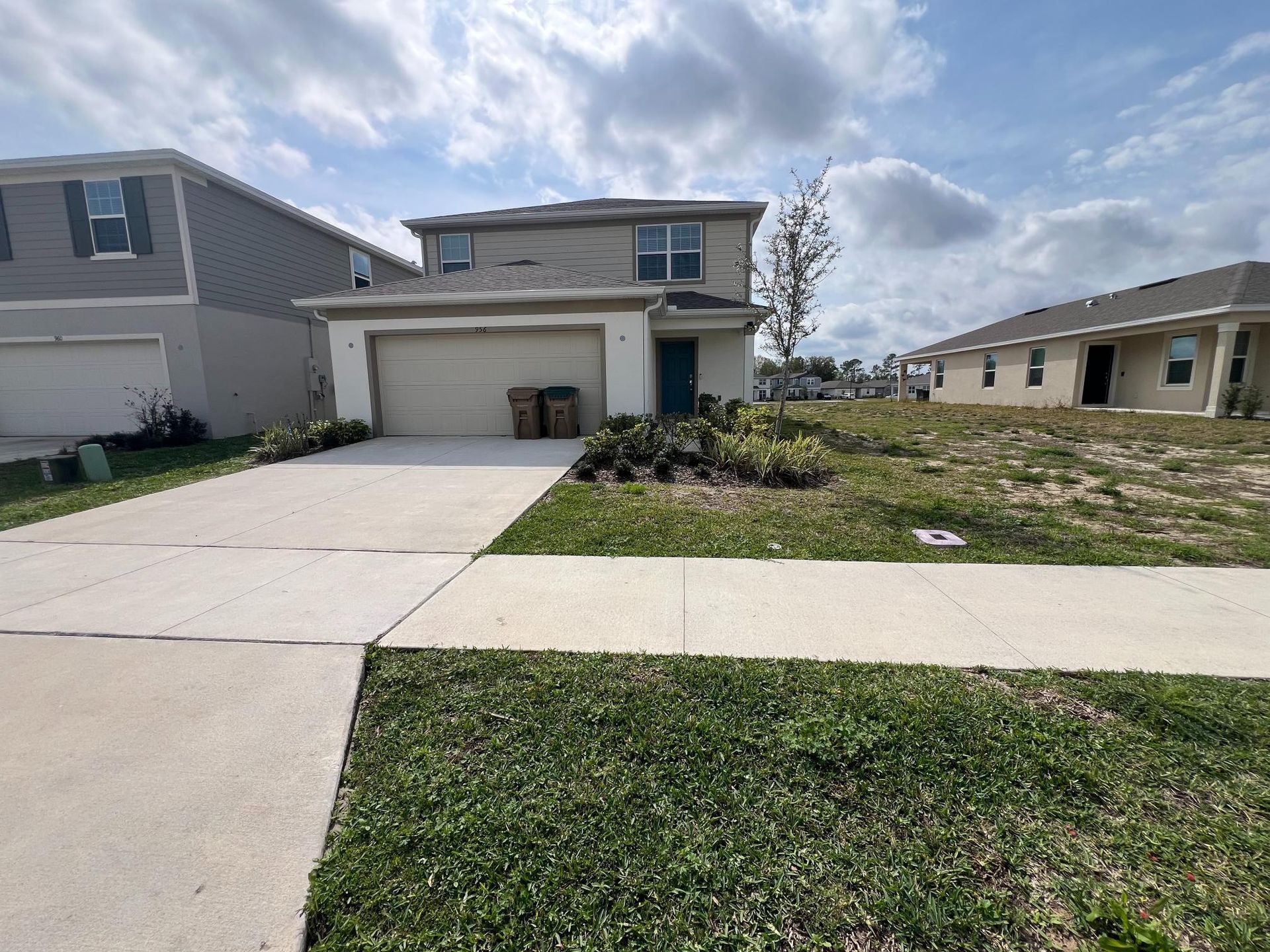 Two-story house with beige siding, driveway, and small front yard with sparse grass under a cloudy sky.