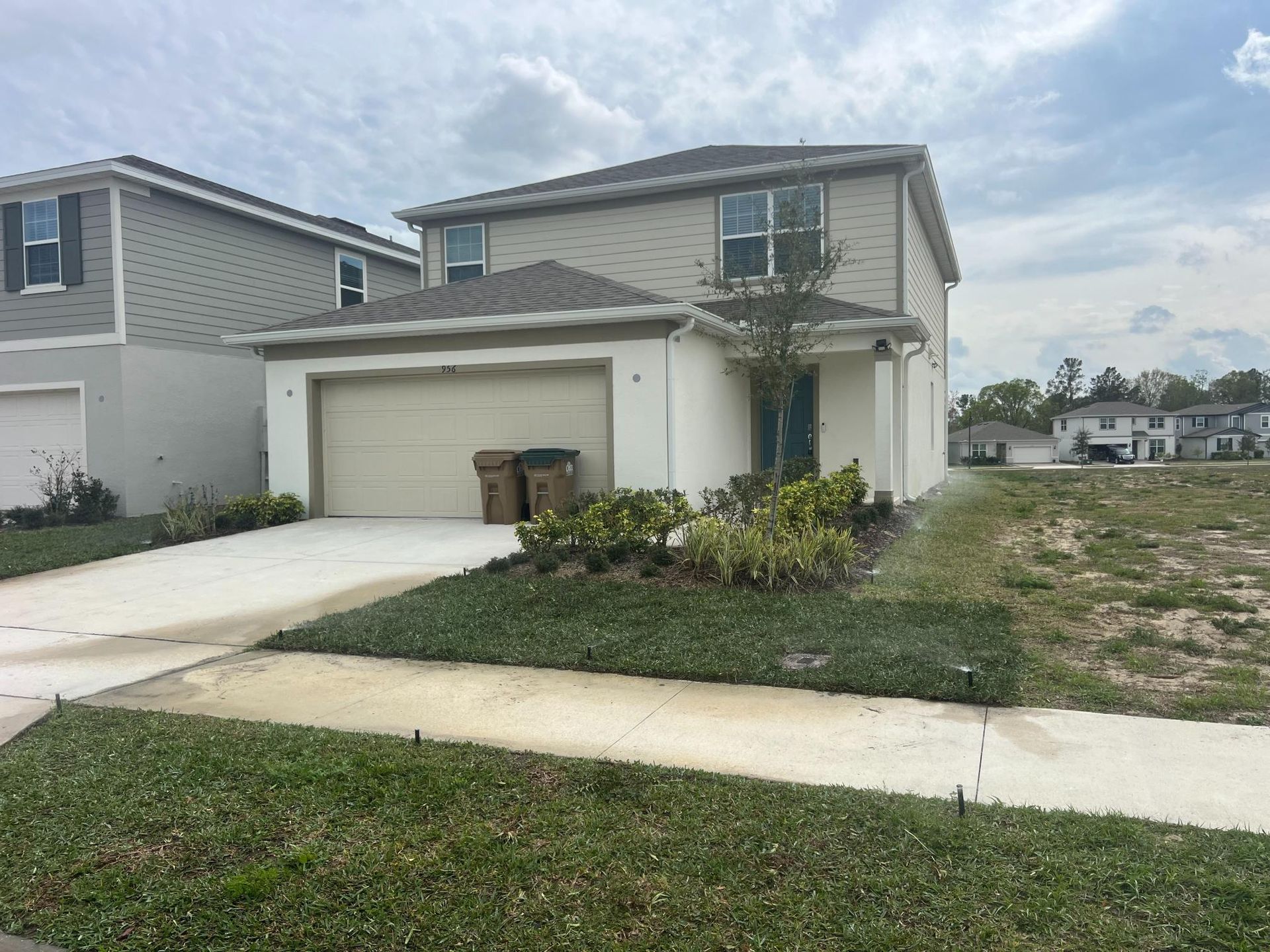 Two-story beige house with a two-car garage, driveway, and patchy grass lawn.