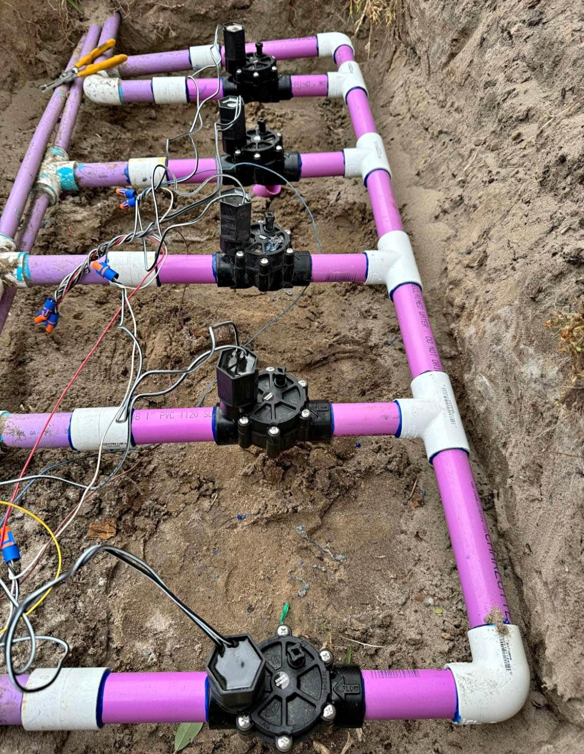 Pink PVC pipes and black irrigation valves installed in a trench. Electrical wires connect the valves.