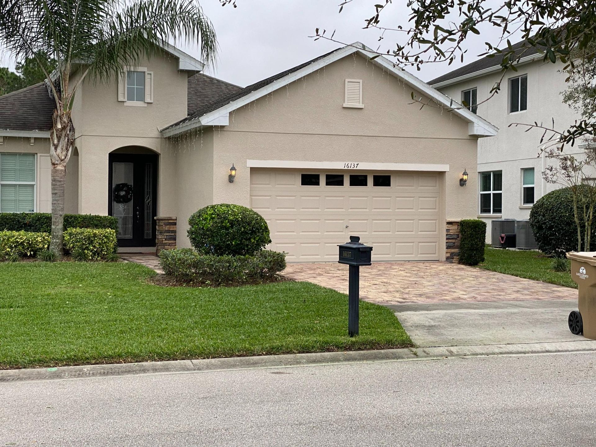 Beige house with brick driveway, green lawn, and trimmed bushes.