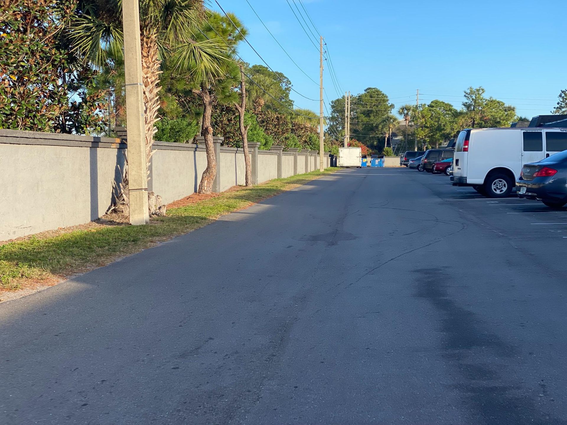 An asphalt road with parked cars and a long concrete wall, under a clear sky.