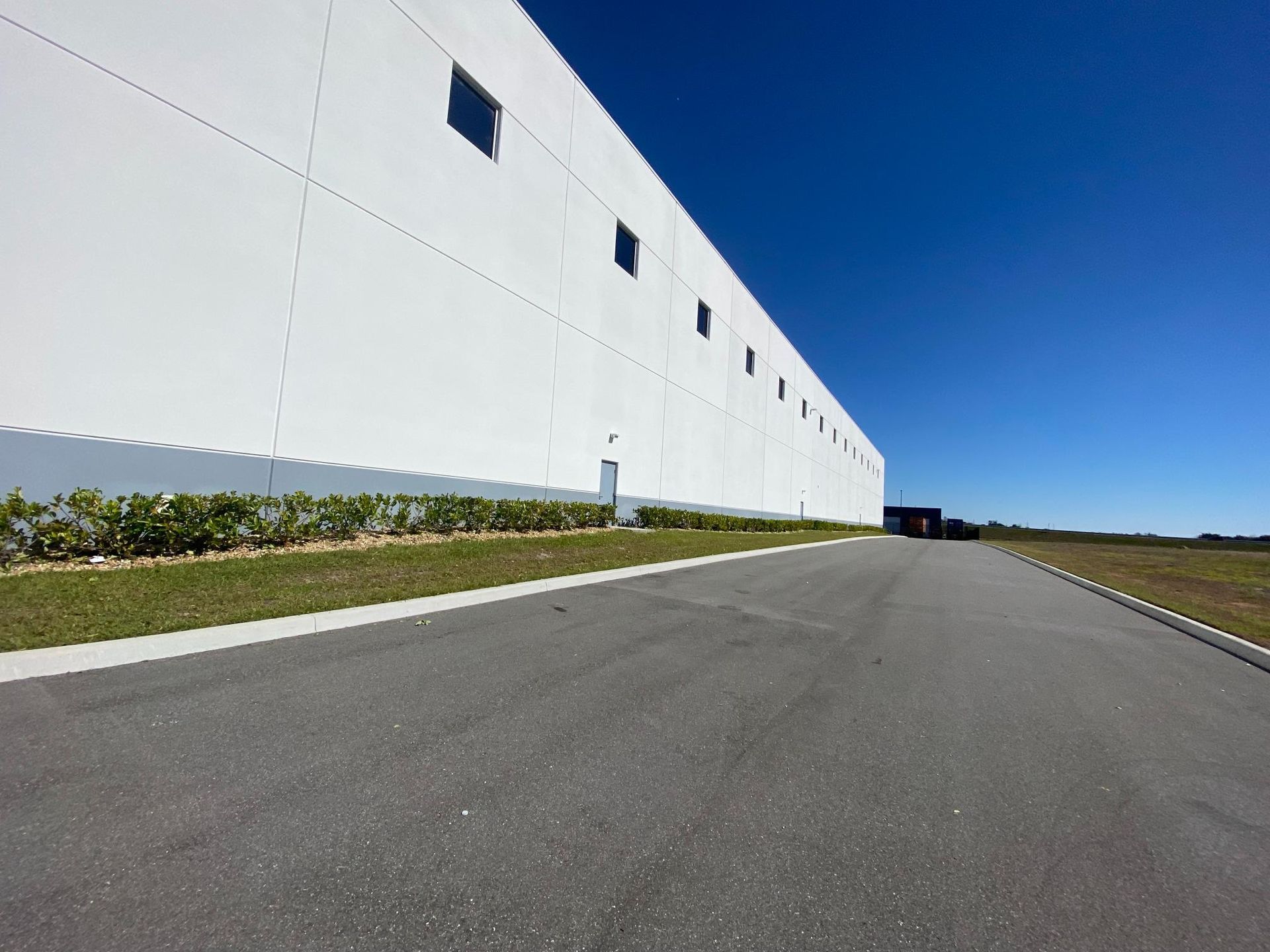 Long, white industrial building with small windows against a clear blue sky, asphalt road.