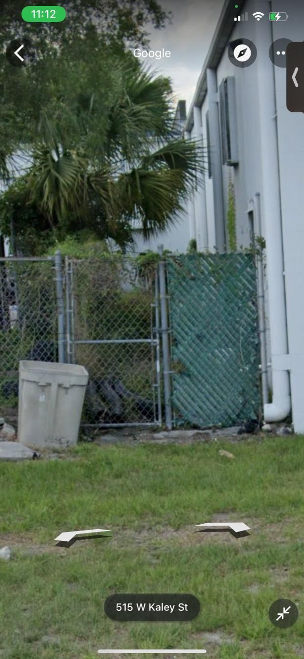 Green chain link fence and gate with trash bin, grassy yard, white building.
