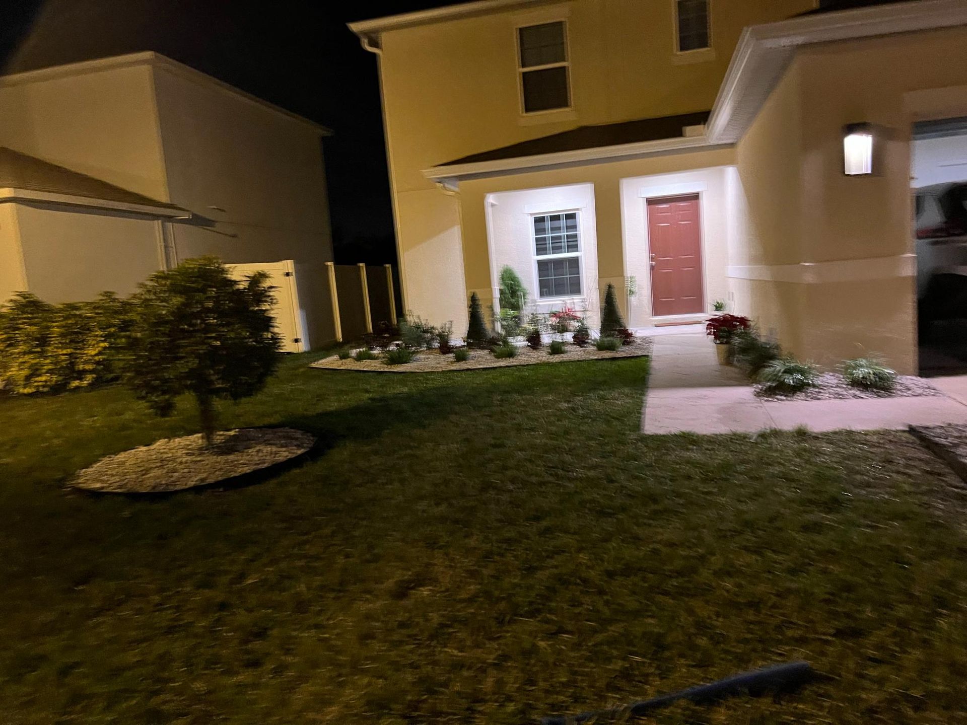 A house at night with a well-lit front yard and landscaping. The front door is red, with lights on.