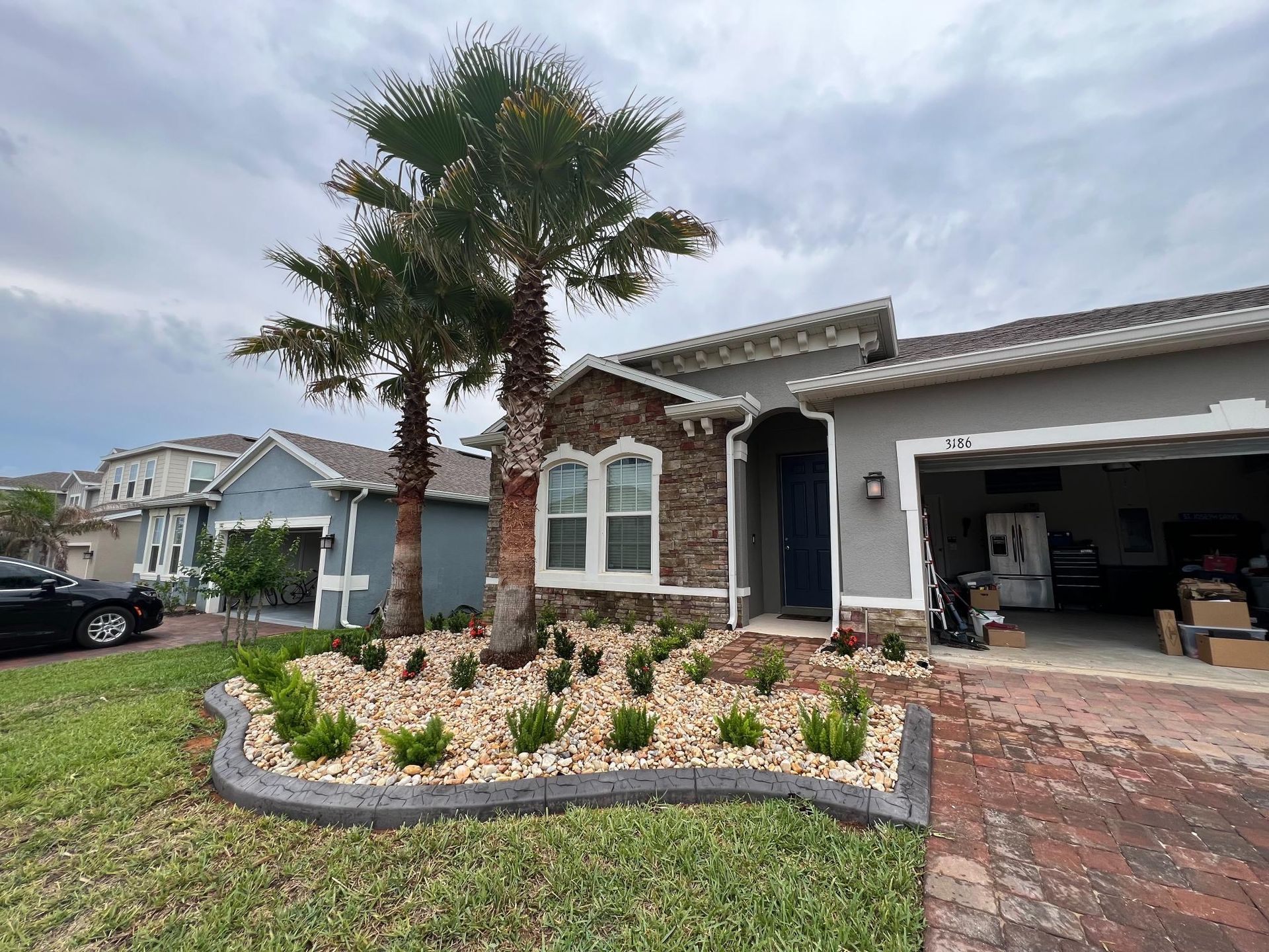 House exterior with palm trees, landscaped garden, gray siding, and a cloudy sky.
