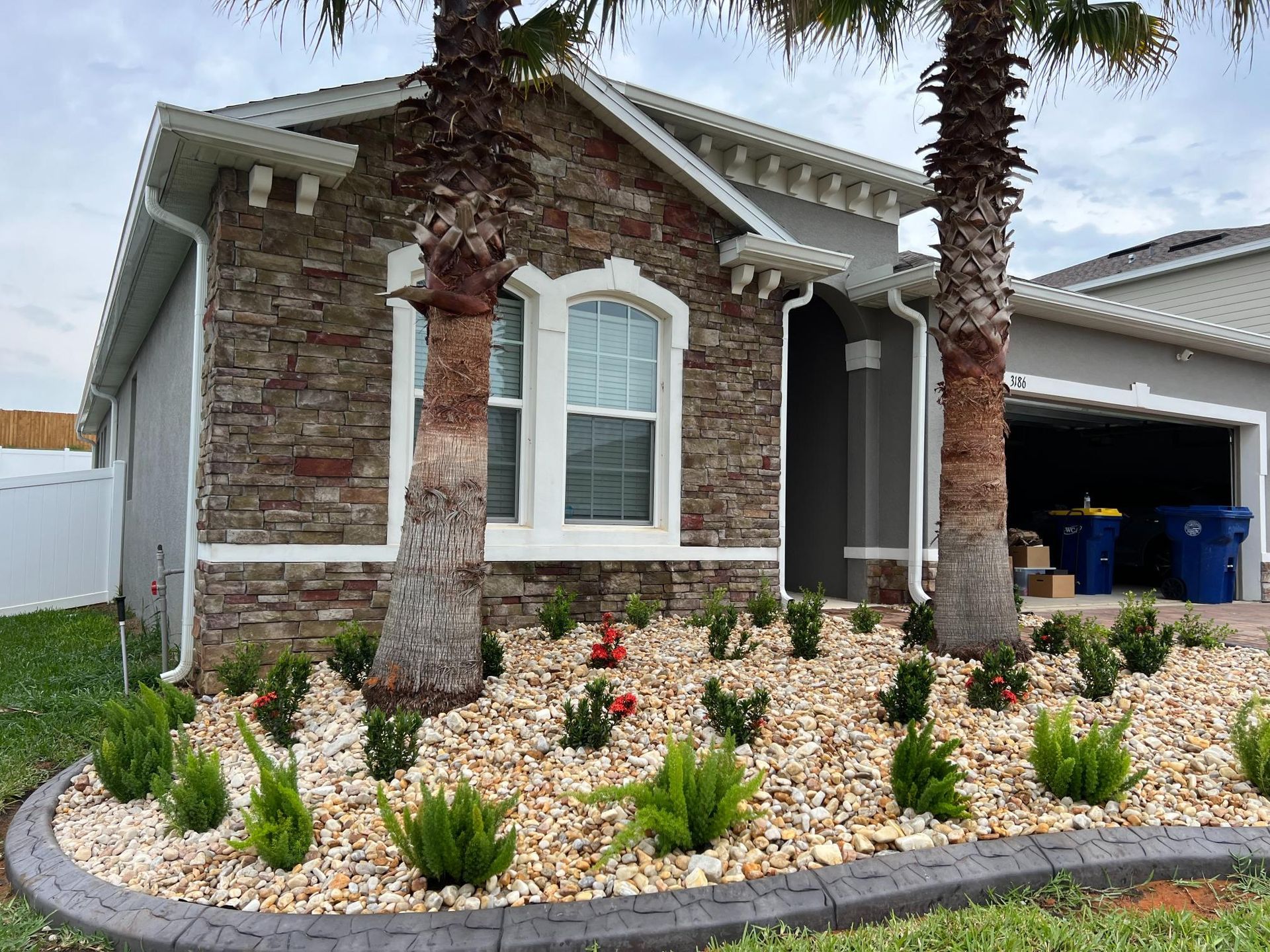 House exterior with stone facade, gray siding, palm trees, and landscaped yard with gravel and plants.
