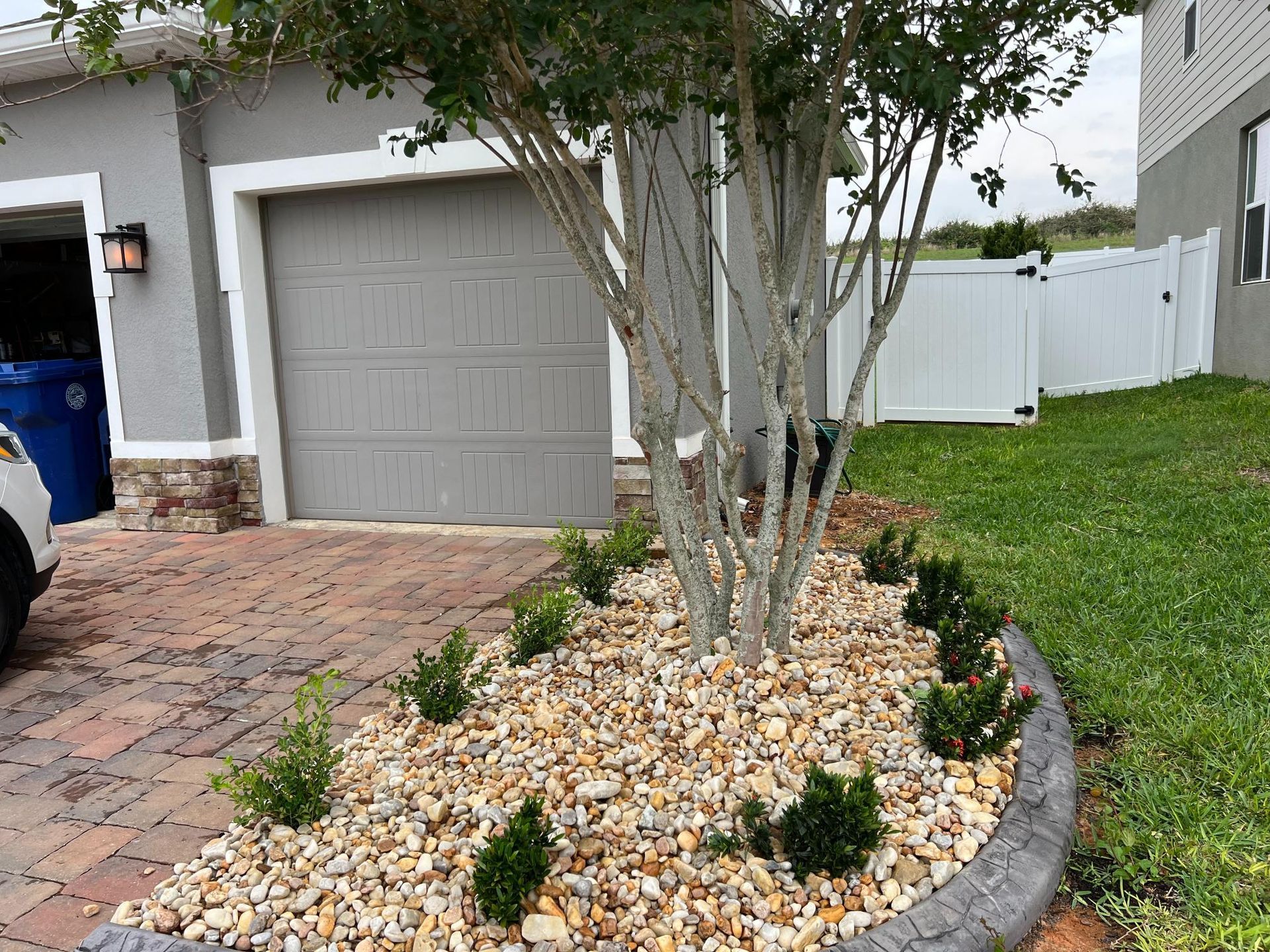 A landscaped front yard with a tree, gravel bed, small green shrubs, and a gray garage door.