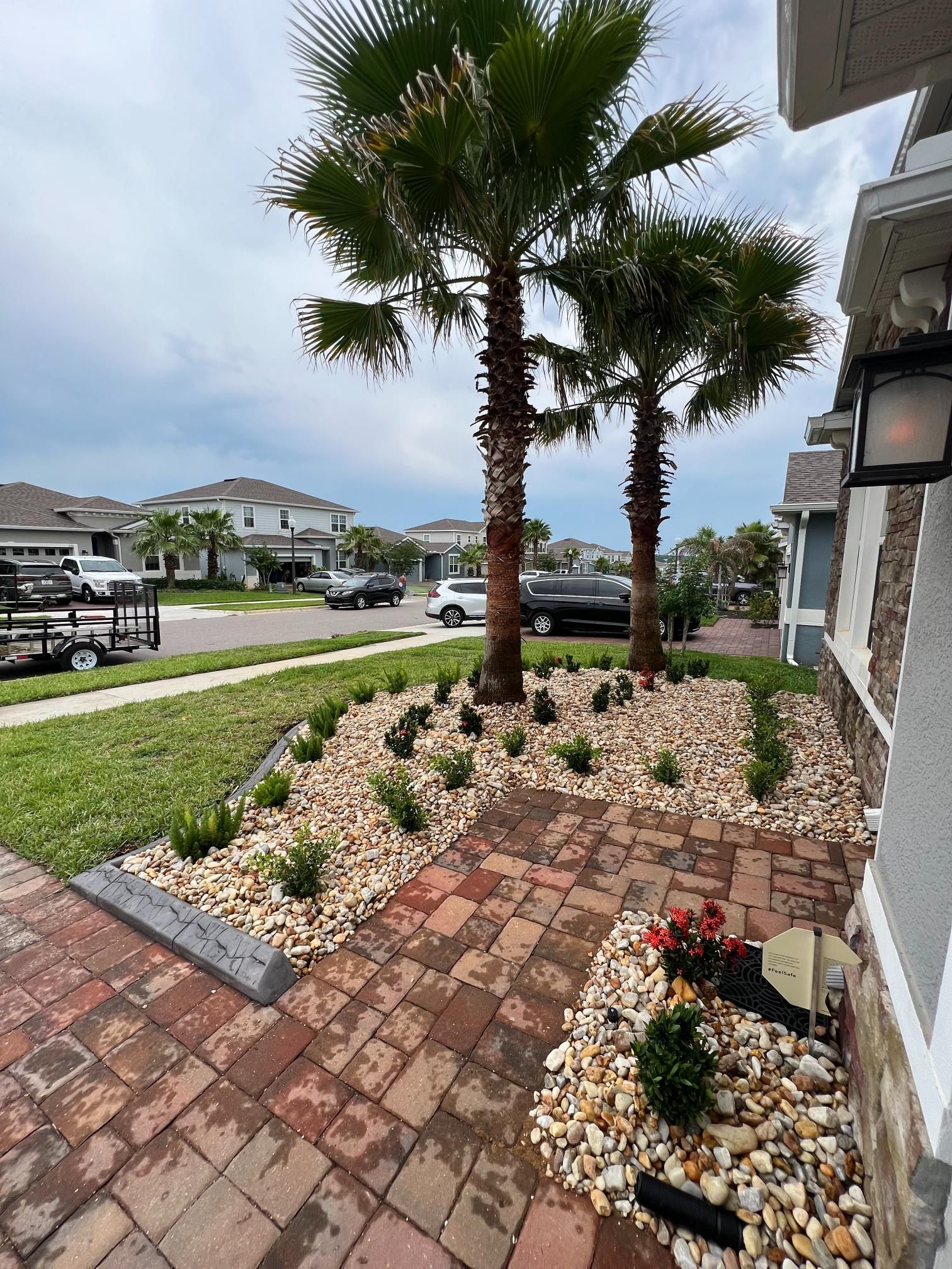 Landscaped yard with palm trees, stone path, flower beds, and houses in the background.