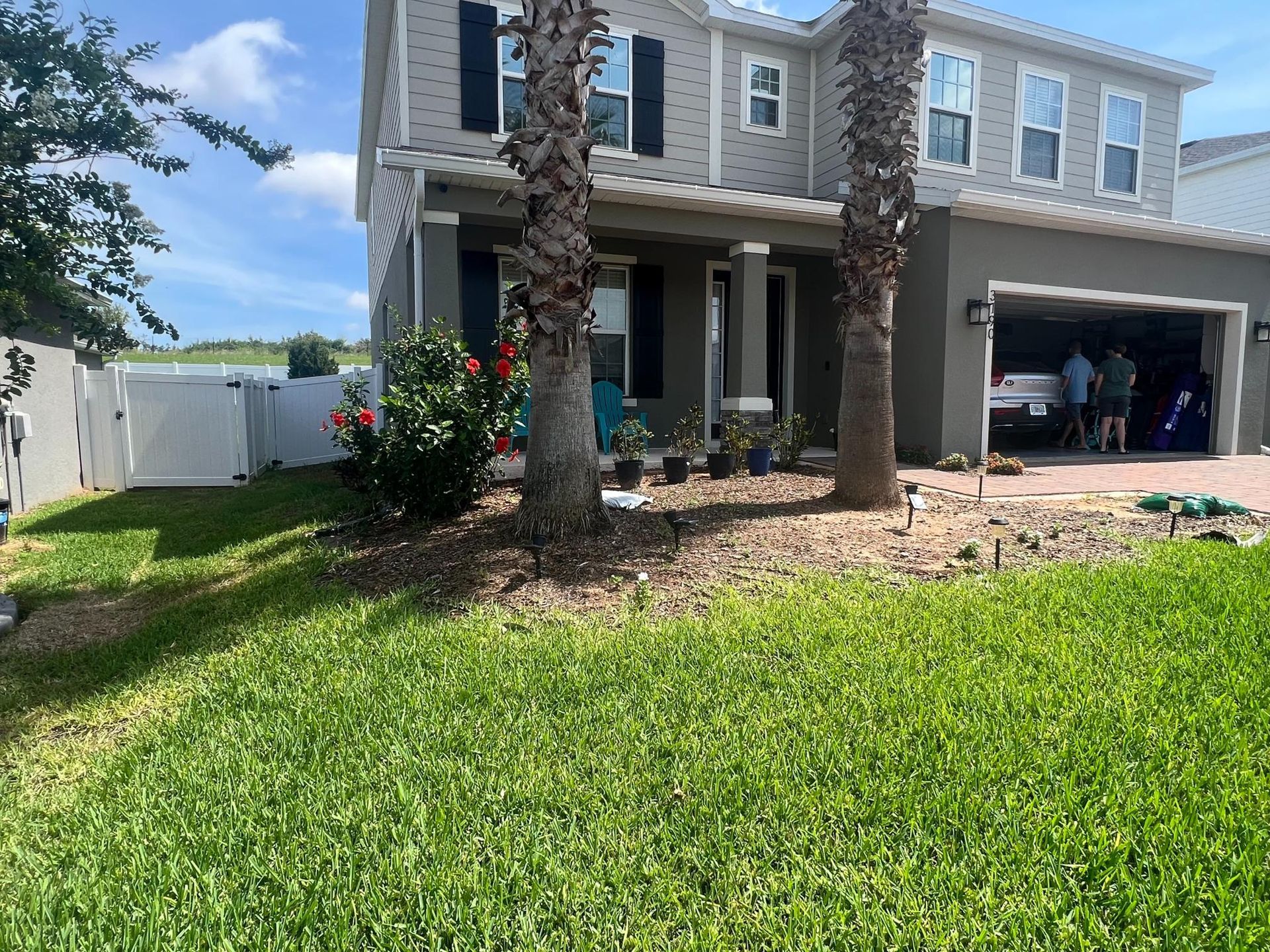 Two-story house with palm trees, garage door open. People visible inside, green lawn. Sunny day.
