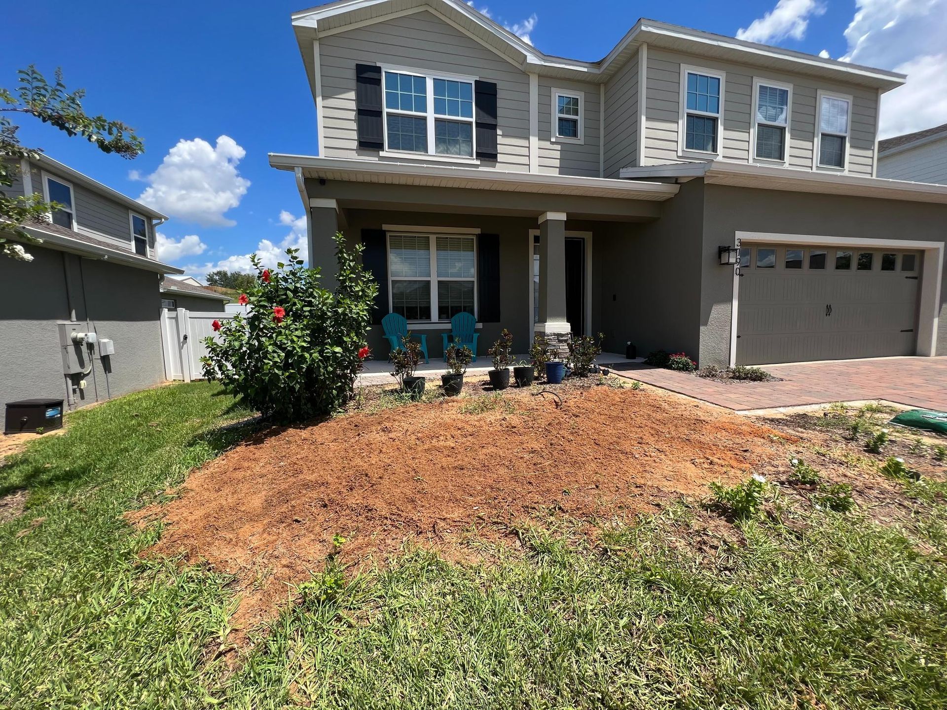 Two-story house with gray siding, red brick driveway, and front yard with mulch and plants. Blue sky.