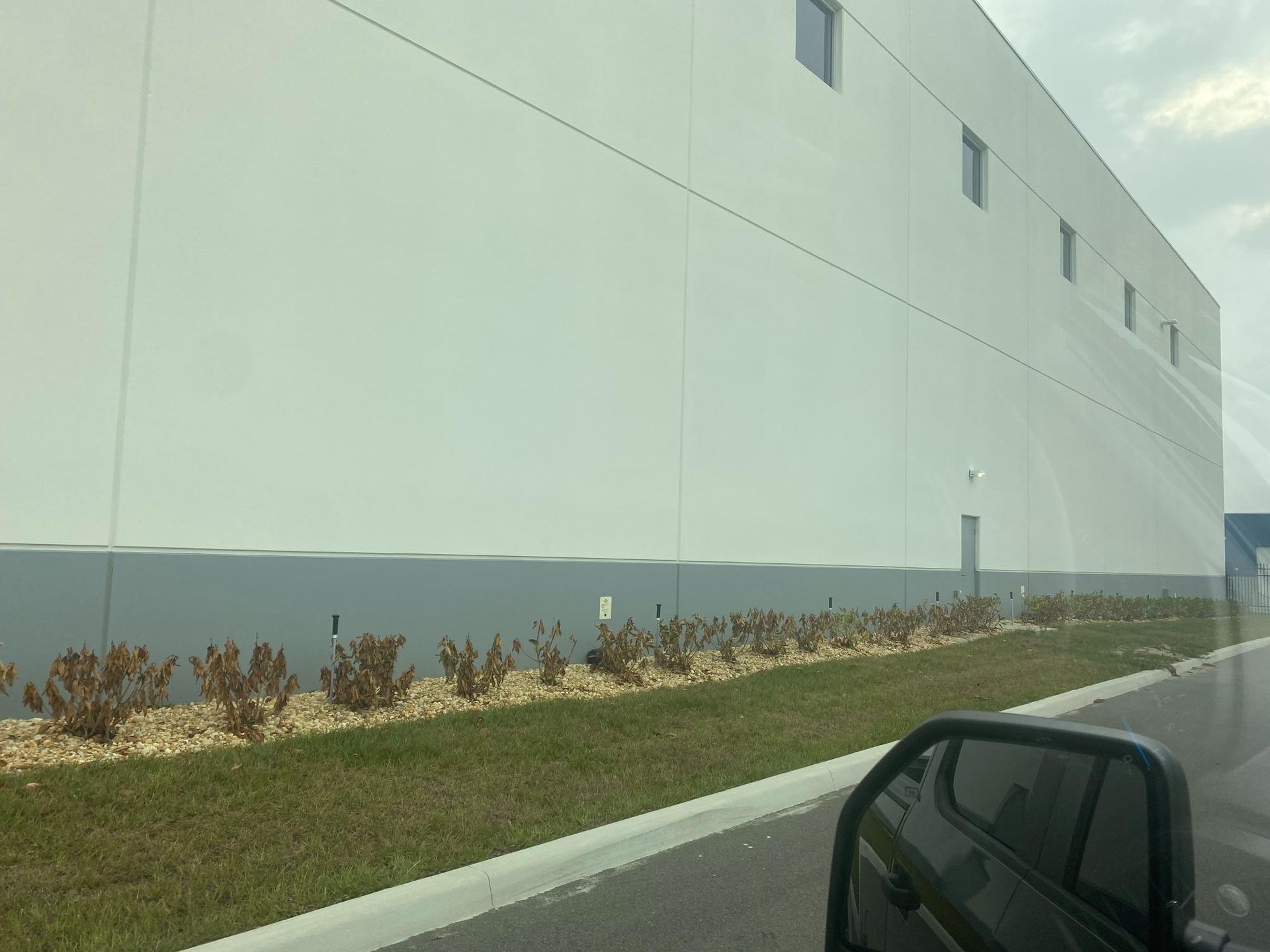 Large white industrial building with a row of dead bushes and grass in front.