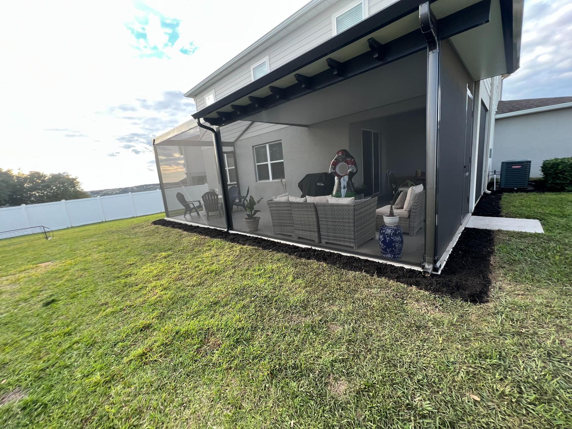 Screened porch with patio furniture on a lawn. Person stands inside porch. Black trim, light colored house, and green grass.