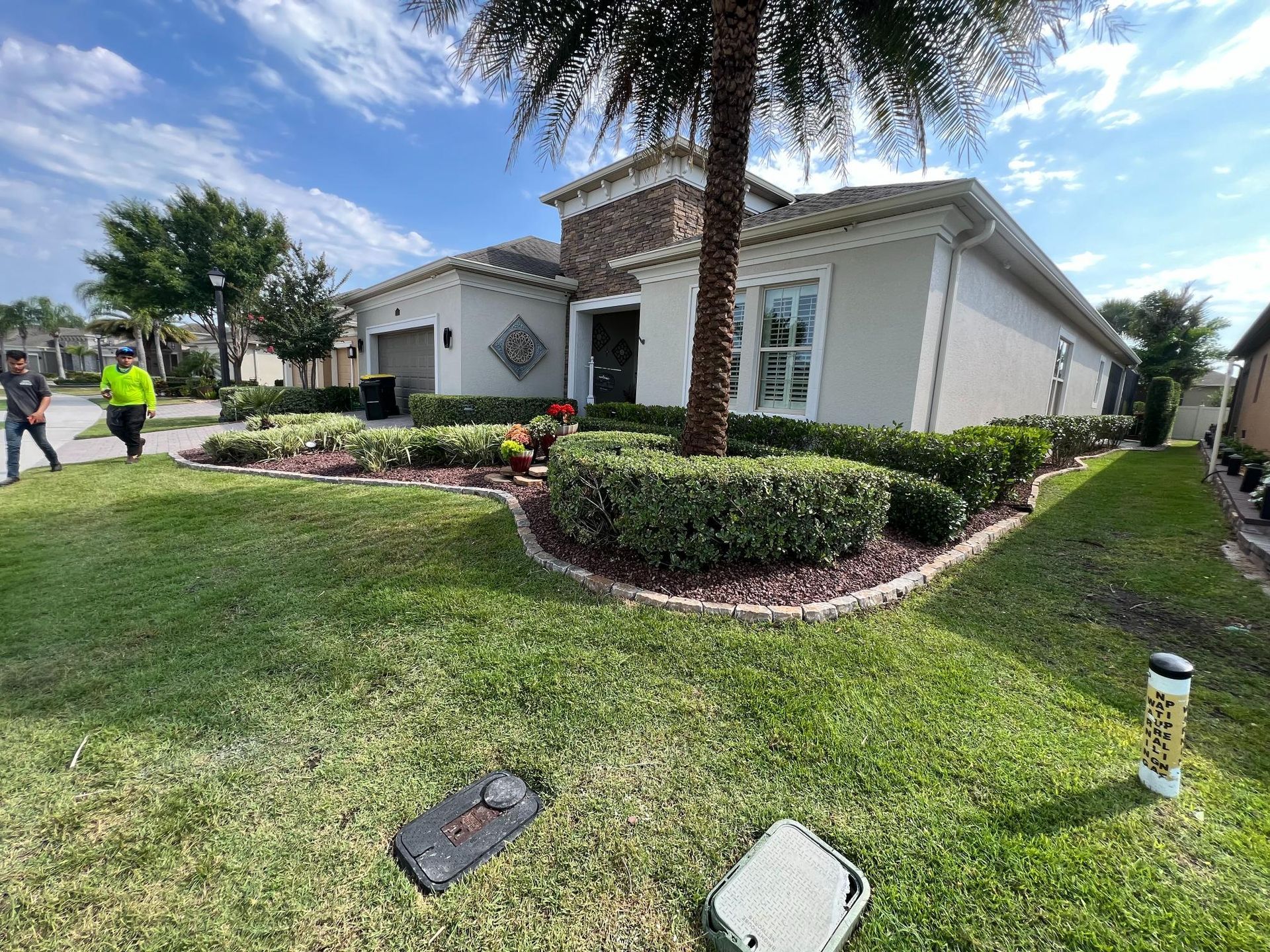 House with lush landscaping, lawn, and a palm tree, with workers in the background.