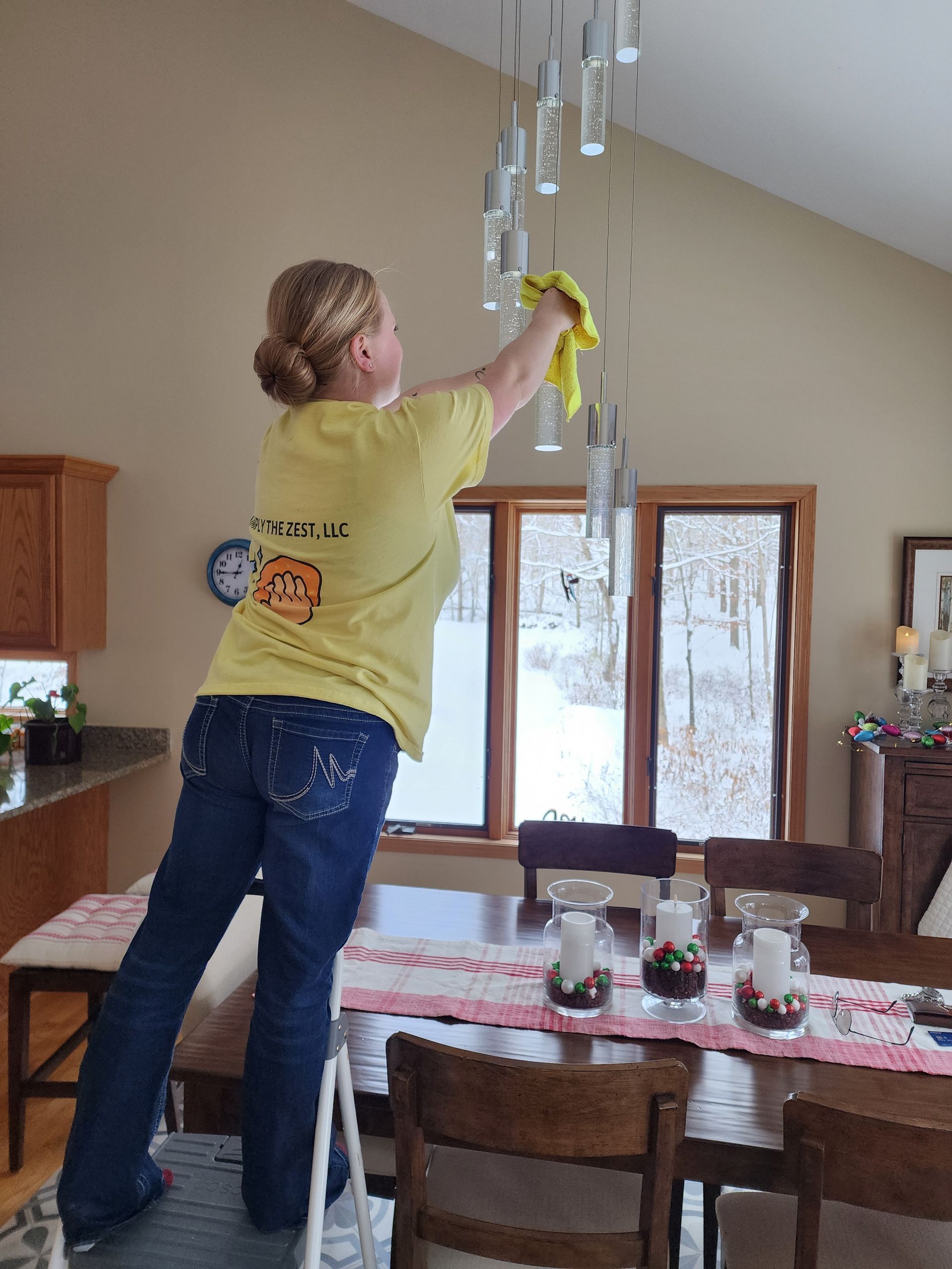 Woman on a step stool dusting a modern chandelier above a dining table near a window.