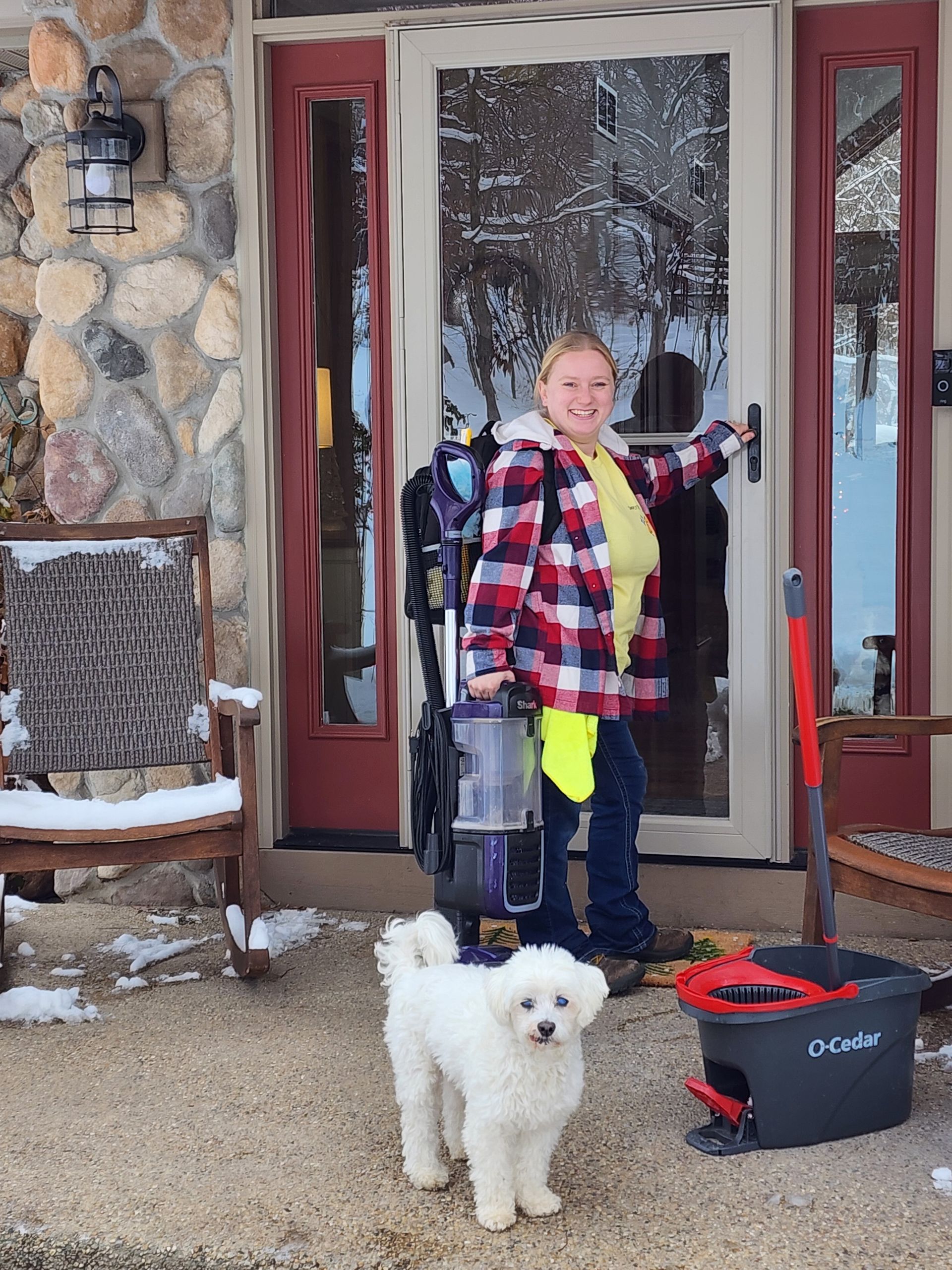 Person in plaid holds cleaning equipment, white dog by their feet, in front of a house entrance.