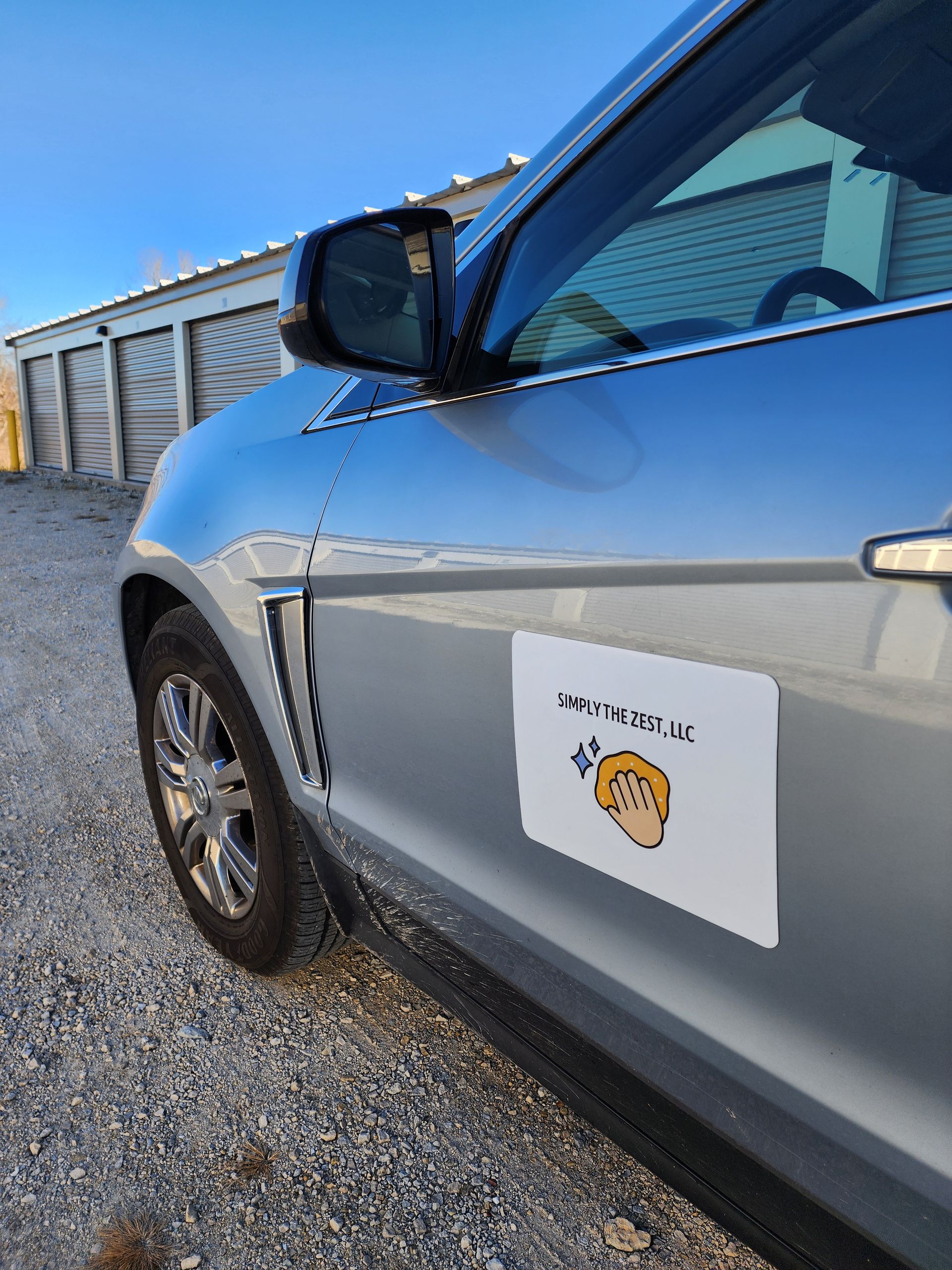 Silver car with a white sign showing a cartoon head and text; parked outside storage units.
