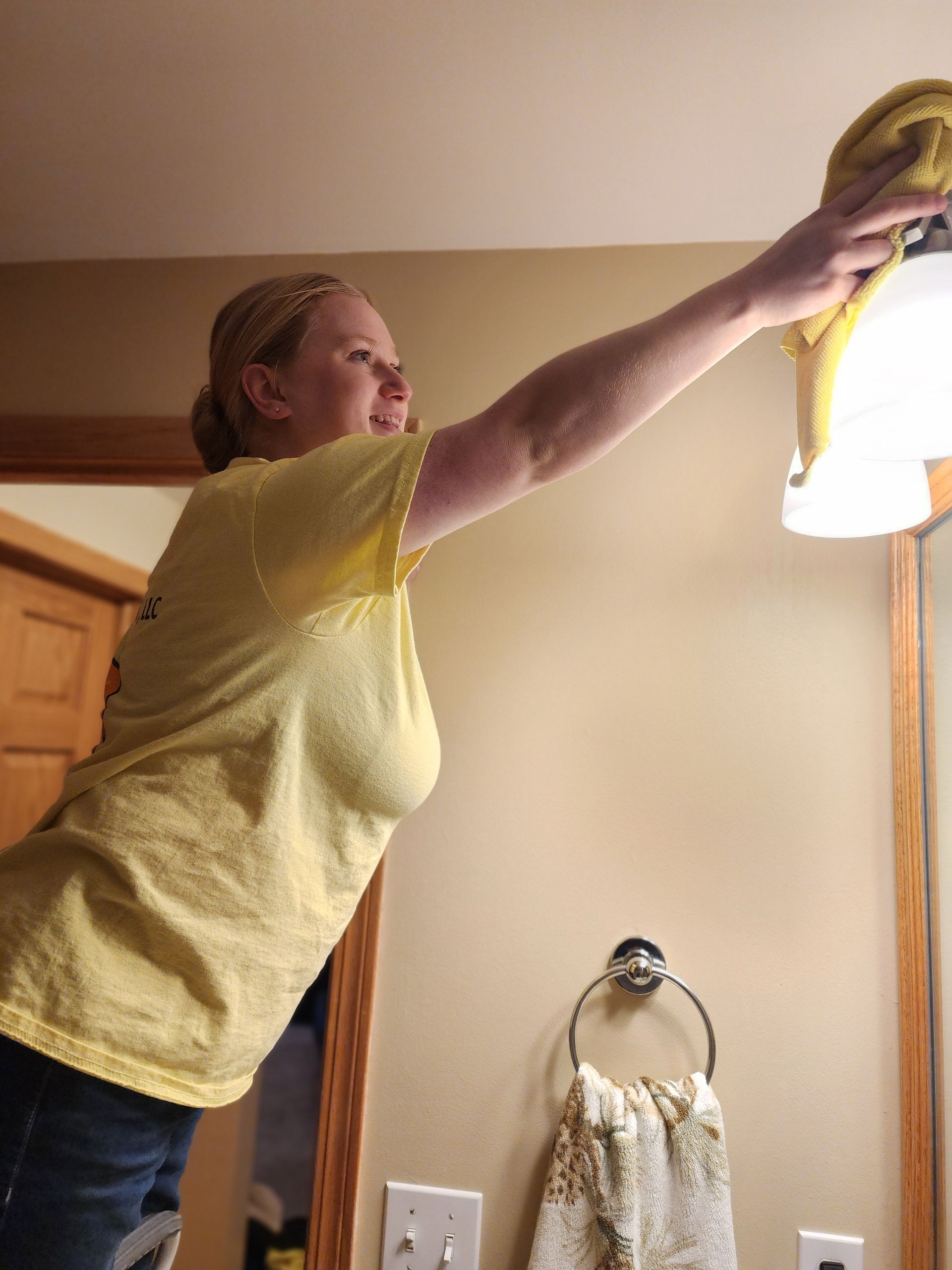Woman in yellow shirt cleaning bathroom light fixture.