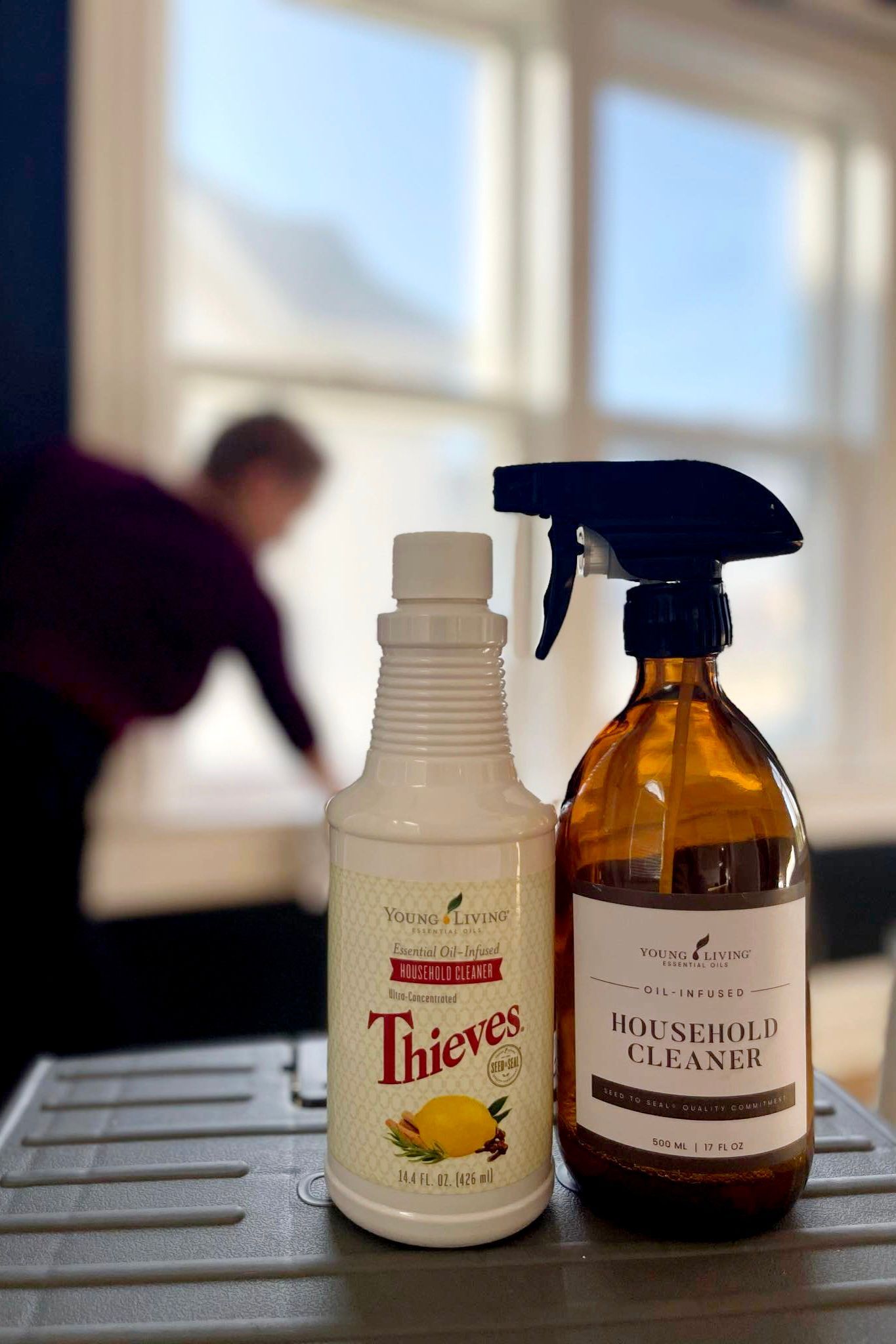 Two cleaning spray bottles in front of a woman cleaning a window.