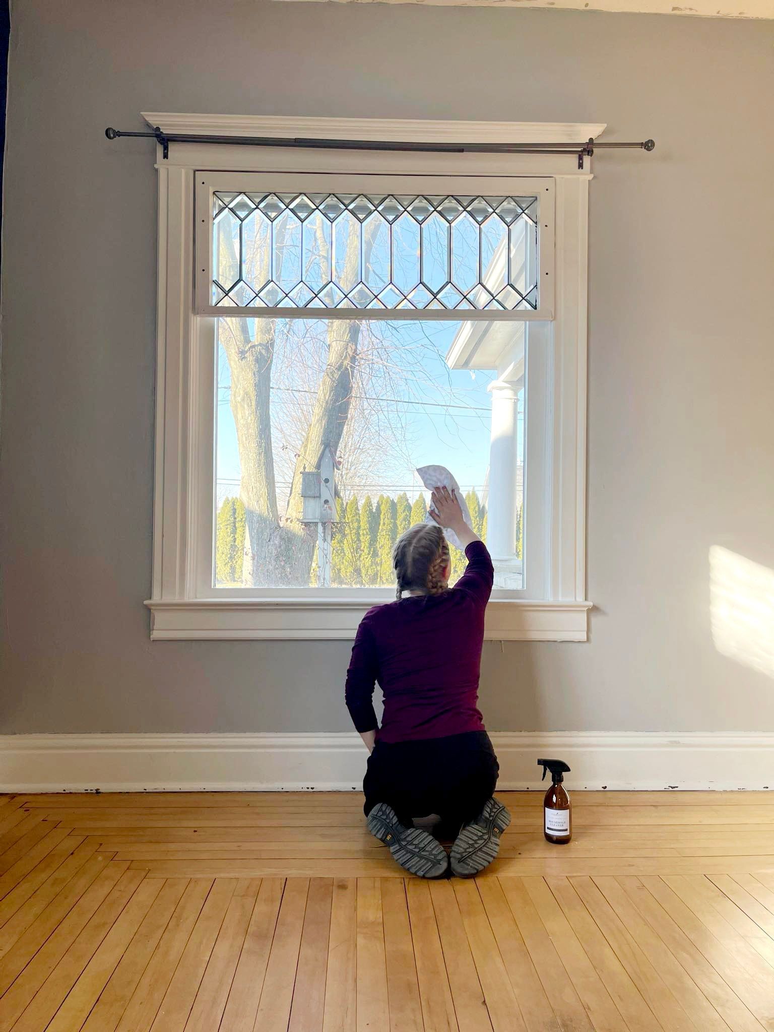 Person kneeling, cleaning a window with a view of trees outside. Brown bottle on floor.