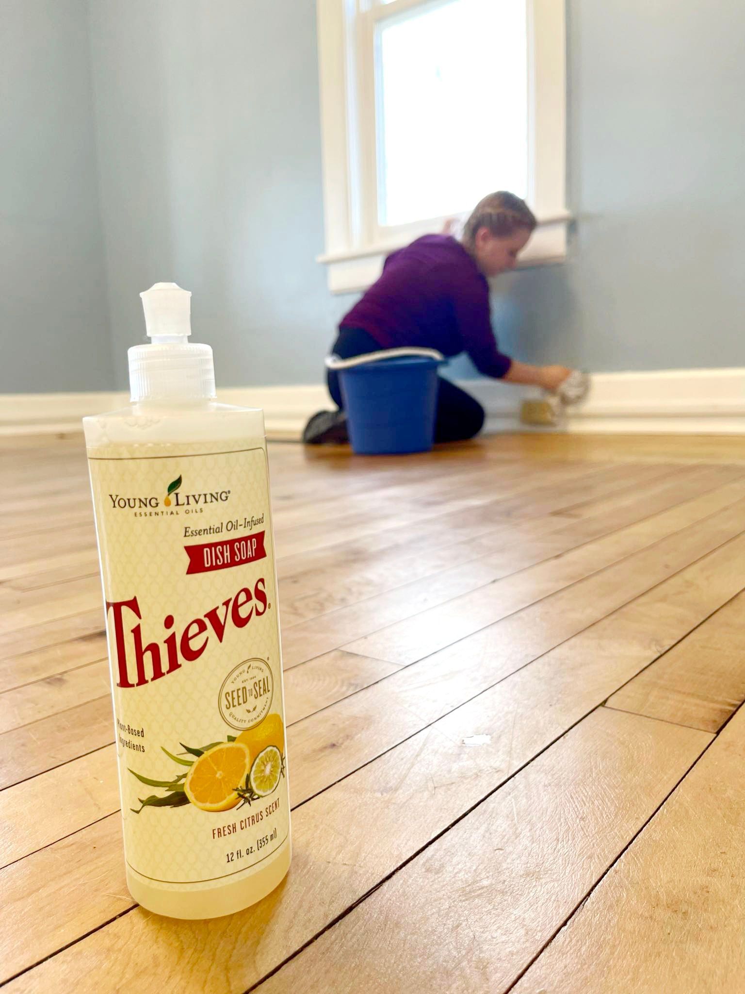 A bottle of Thieves cleaner on wood floor; a woman cleans baseboards in a room with blue walls.
