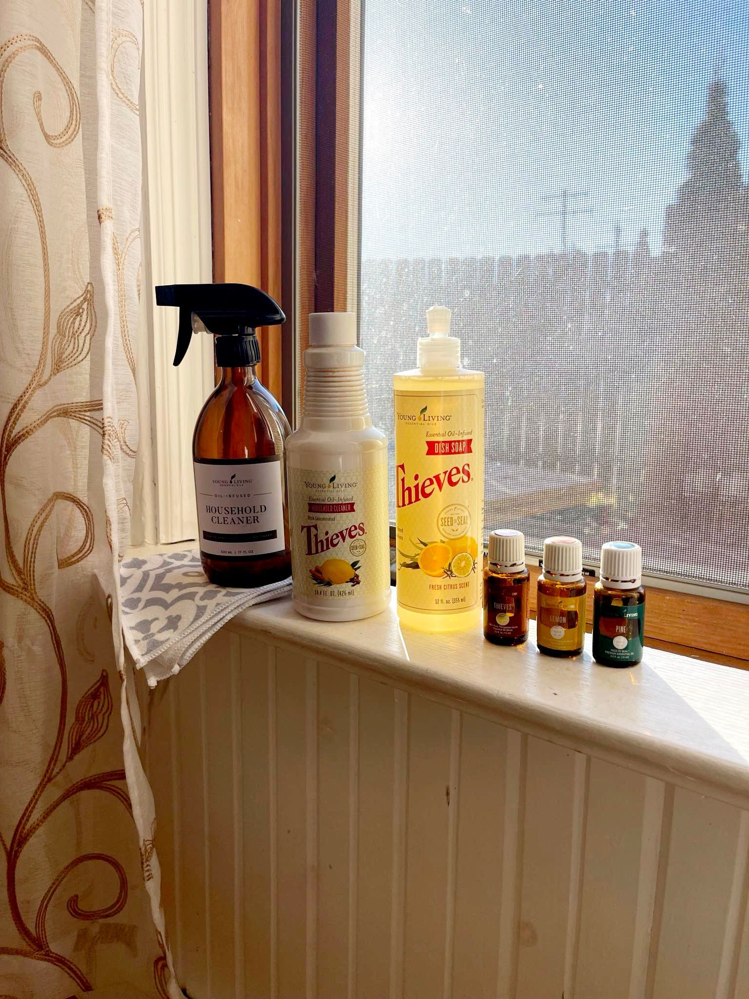 Bottles of cleaning supplies and essential oils on a windowsill next to a window with a screen.