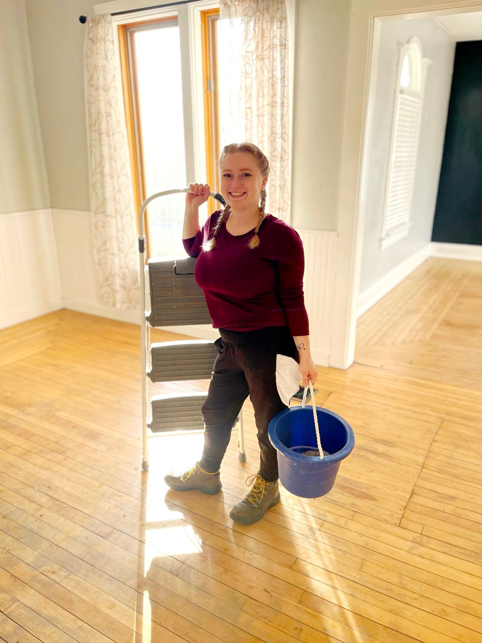 Woman with braids holding a bucket and leaning on a stepladder in a room with hardwood floors.