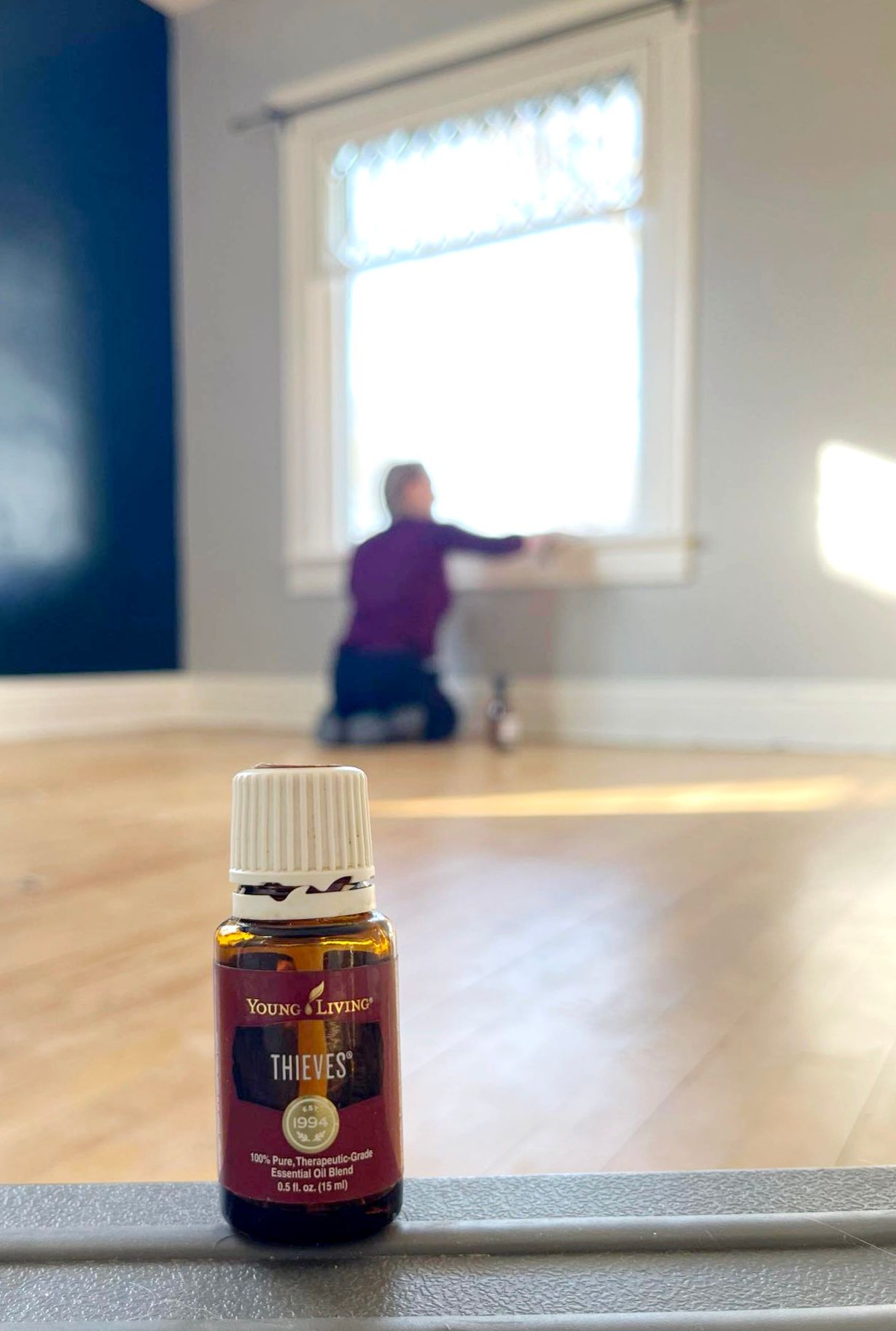 A bottle of Thieves essential oil in front of an empty room. A person is cleaning a window in the background.
