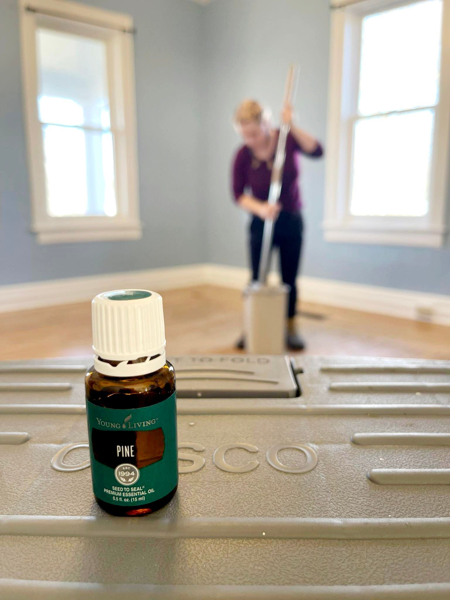 A bottle of essential oil on a box, with a person mopping a room in the background.