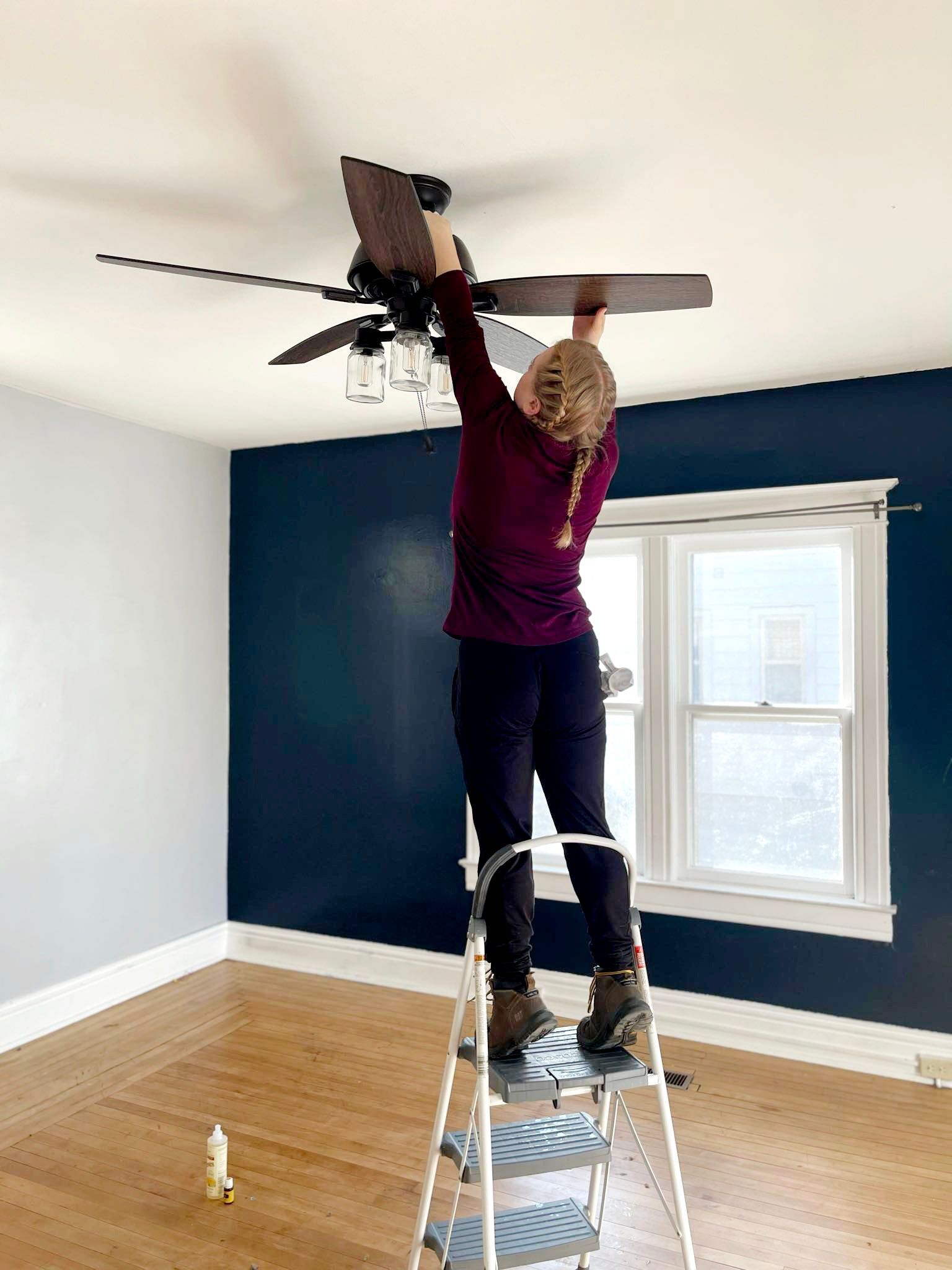 Woman on a step ladder, installing a ceiling fan in a room with blue walls and wood floors.